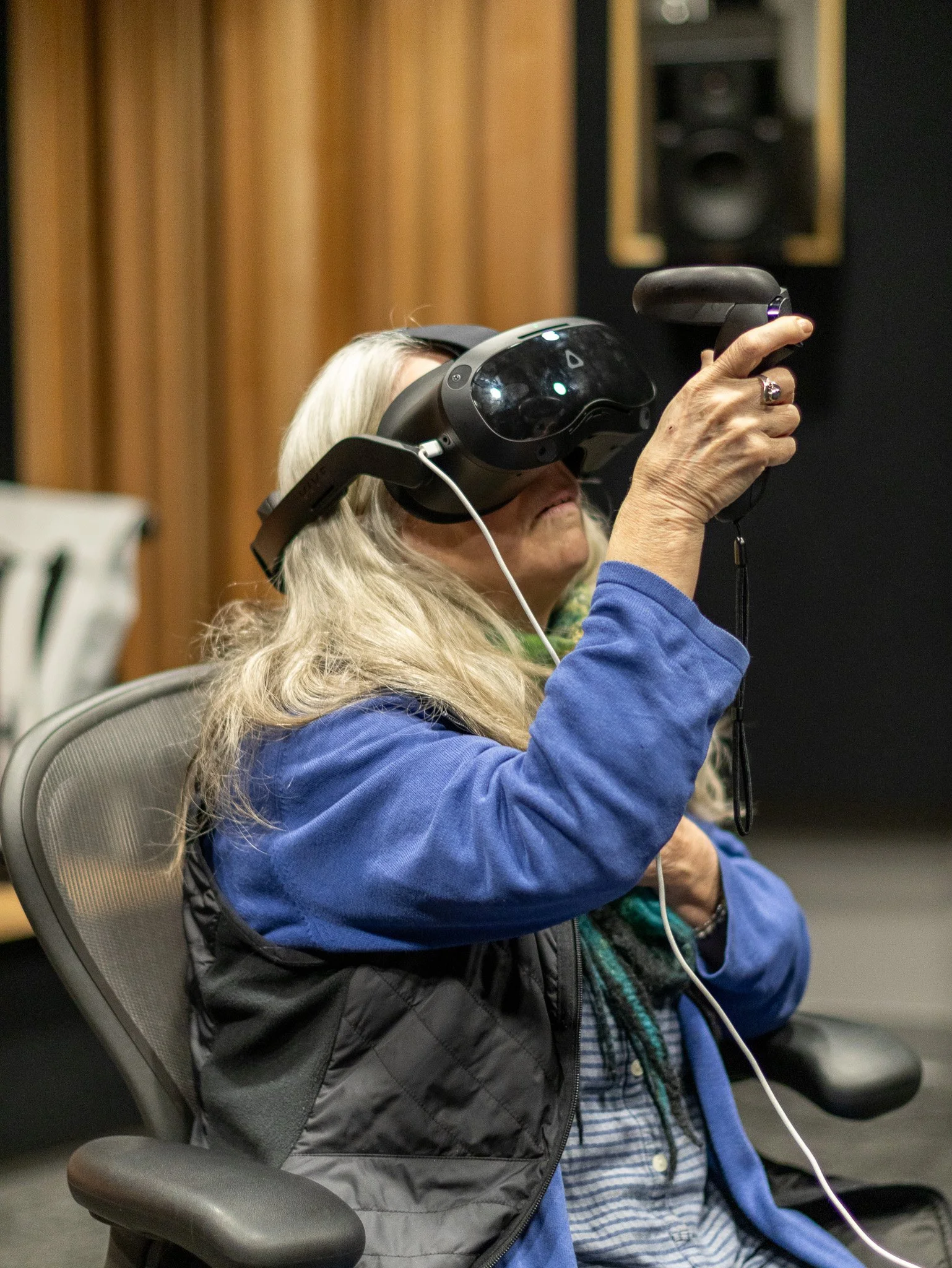 An elderly woman with long, gray hair wearing a blue jacket, plaid shirt, and colorful scarf, sitting in a chair, using a virtual reality headset with headphones, and holding a controller in her right hand.