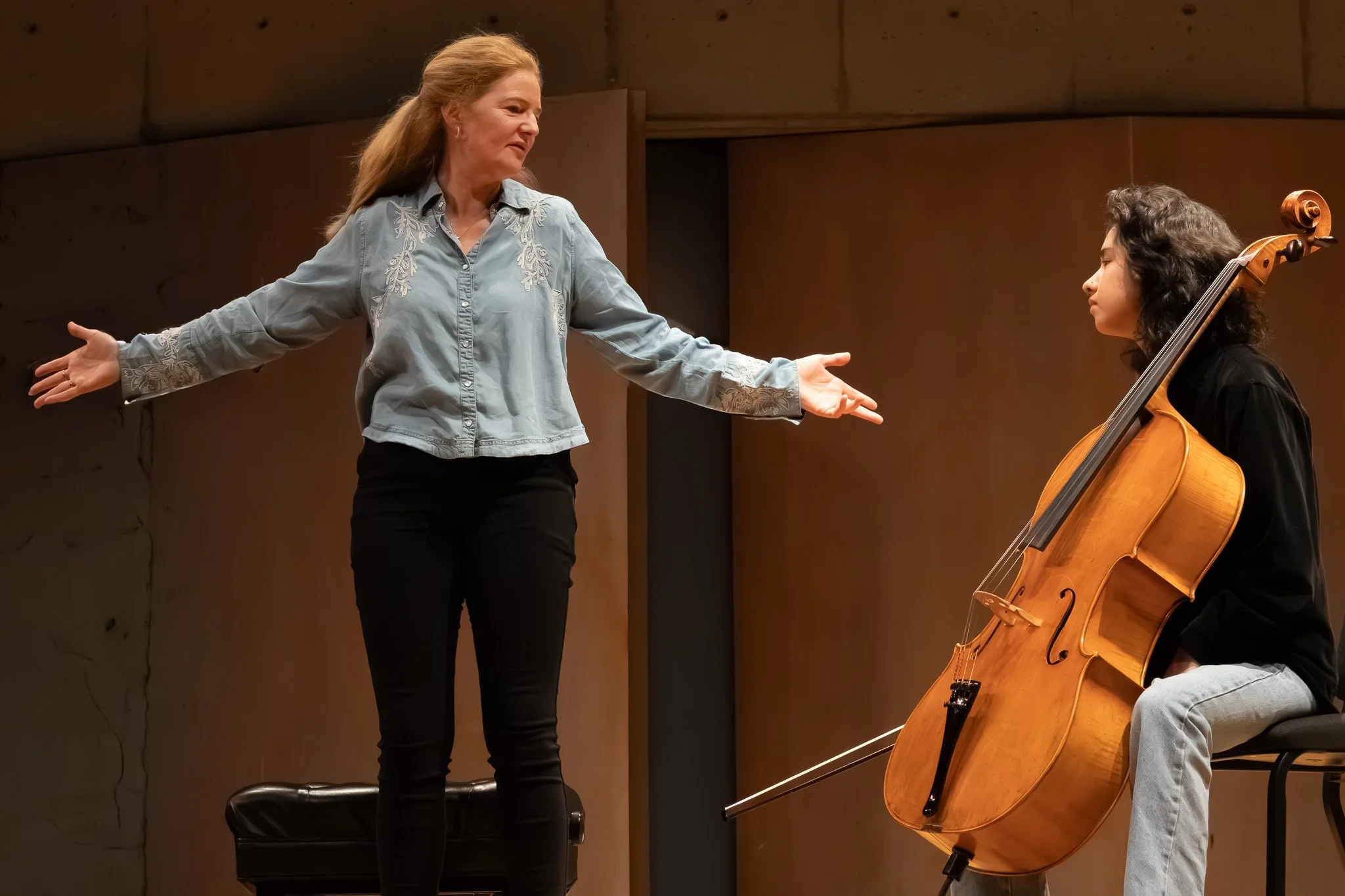 A woman with long, reddish hair wearing a light blue embroidered shirt and black pants is standing on stage with arms outstretched, facing a young woman who is seated and playing a cello. The setting appears to be a concert hall or rehearsal space.