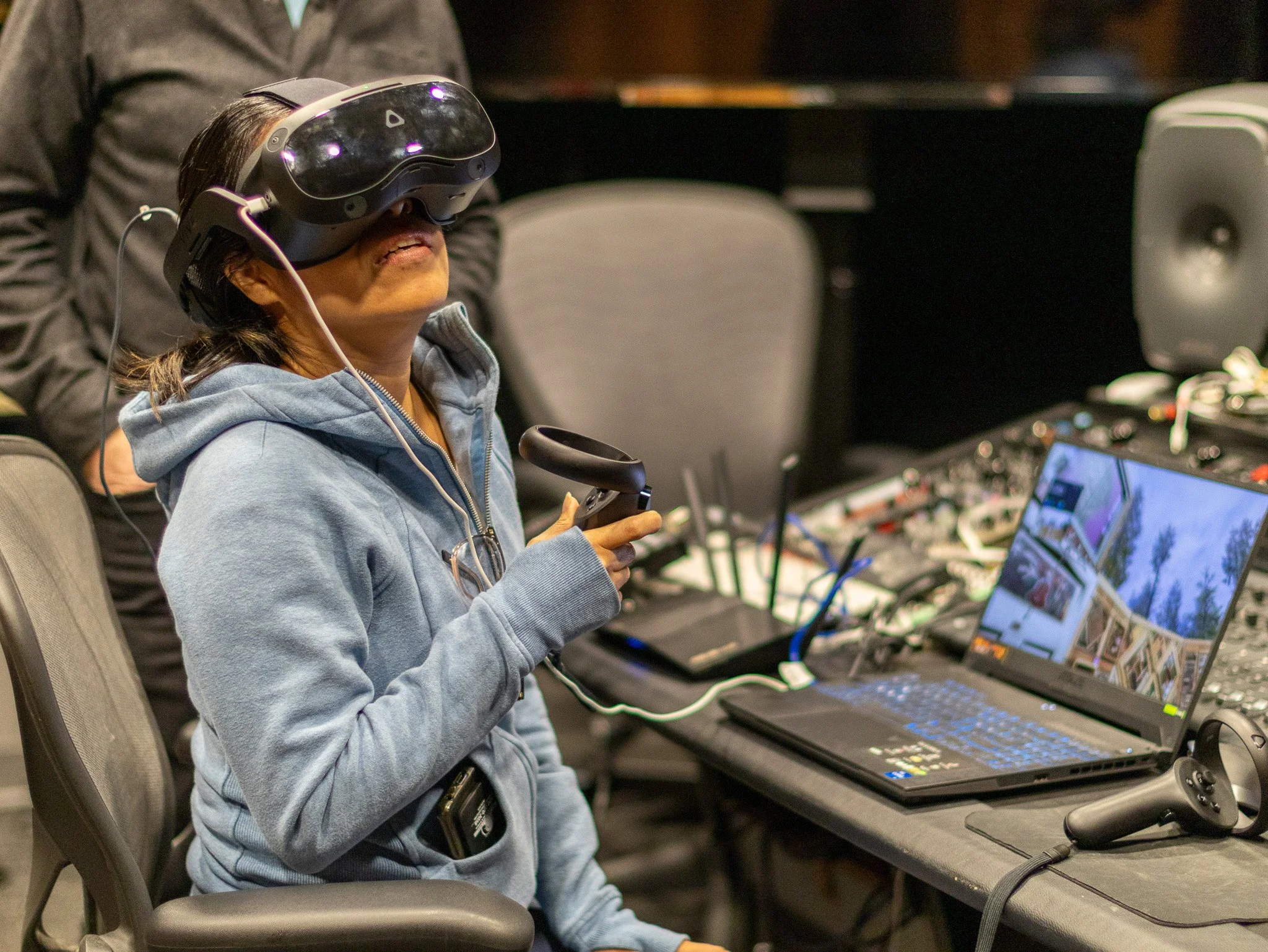 A woman in a gray hoodie and glasses hanging from her shirt is sitting at a desk with multiple laptops, using a virtual reality headset and holding a small controller.