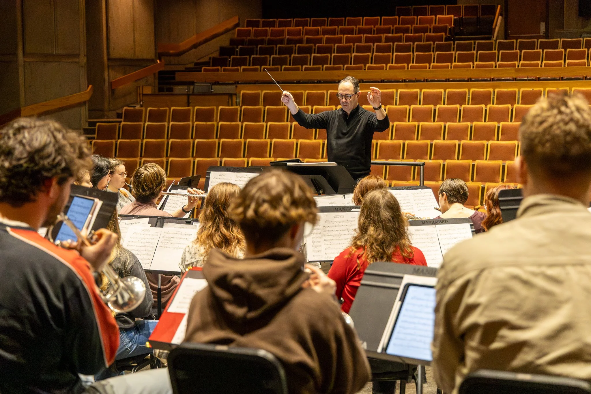 Orchestra conductor leading musicians in a large auditorium with orange seats.