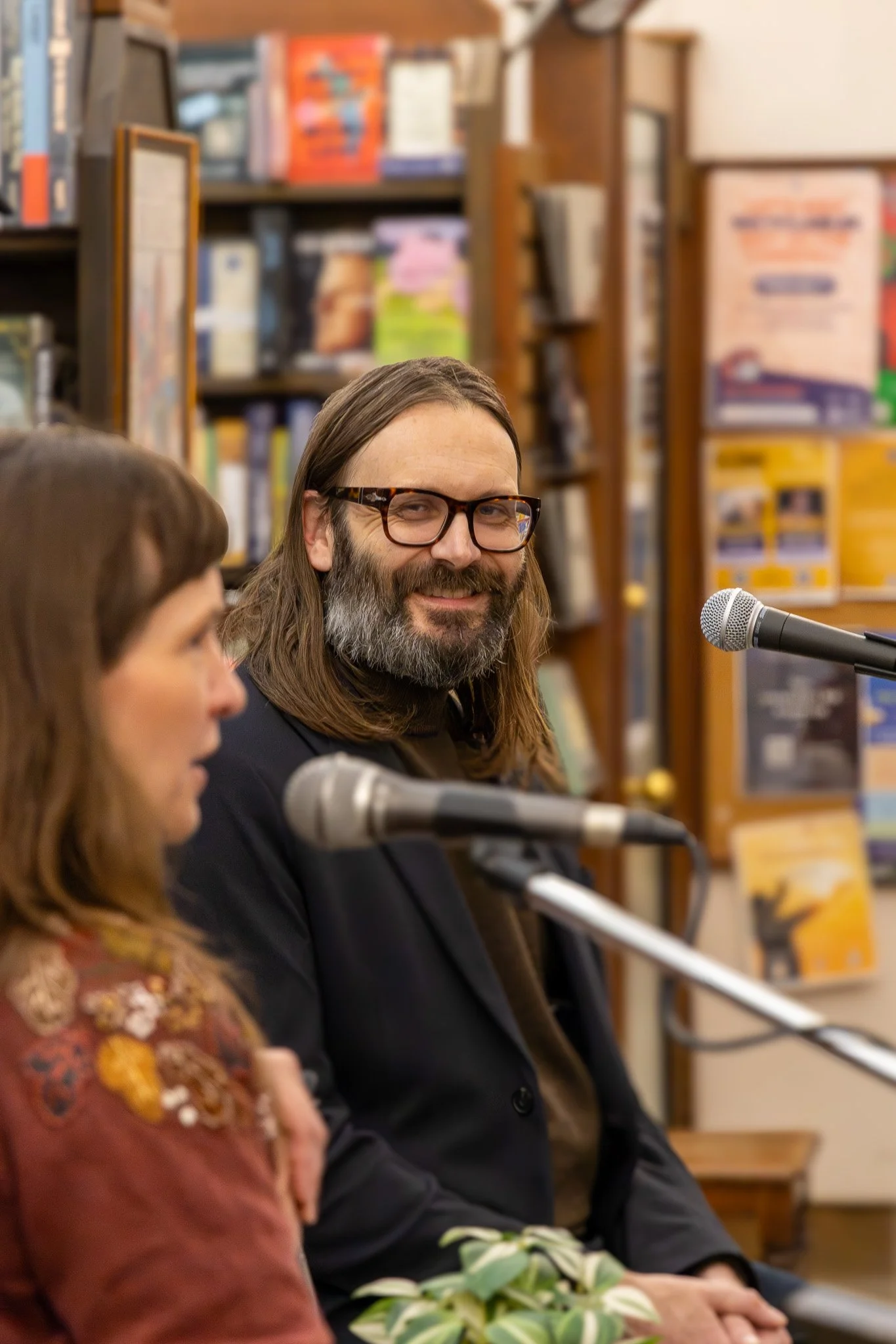 A man with long hair, glasses, and a beard, smiling, sitting at a table with two microphones, with bookshelves in the background.