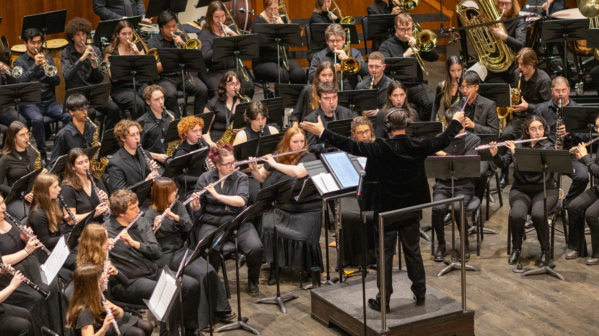 An orchestra performing on stage, with musicians playing wind, brass, and percussion instruments, directed by a conductor.