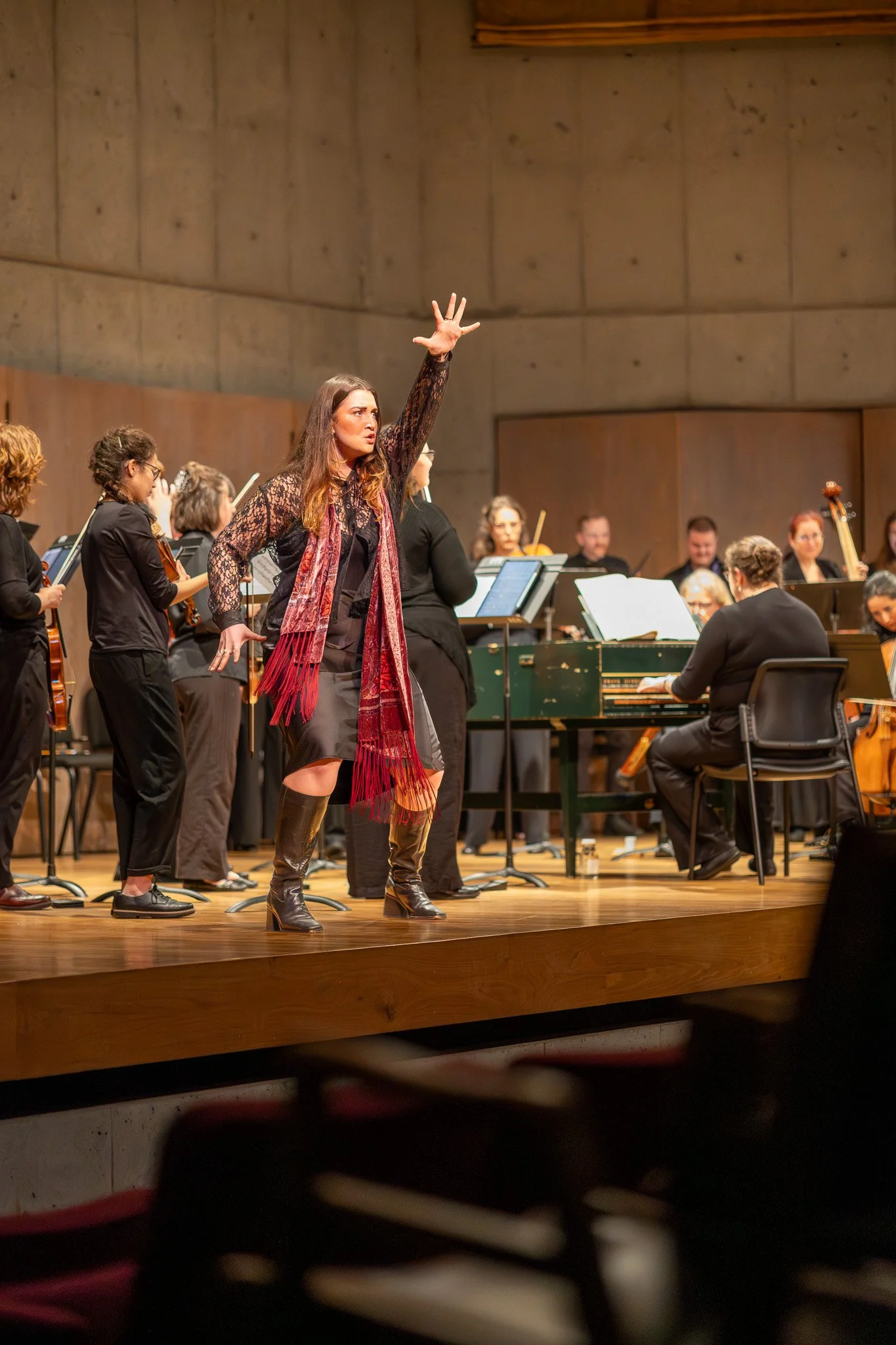 A female conductor with long brown hair, wearing a black lace top, red scarf, and leather boots, leading an orchestra in a concert hall with a wooden stage.