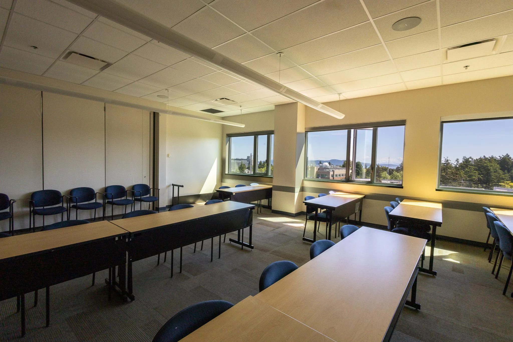 An empty classroom with beige walls, large windows showing a cityscape and greenery, rows of blue chairs against the wall, and light-colored tables with black frames, illuminated by natural sunlight.