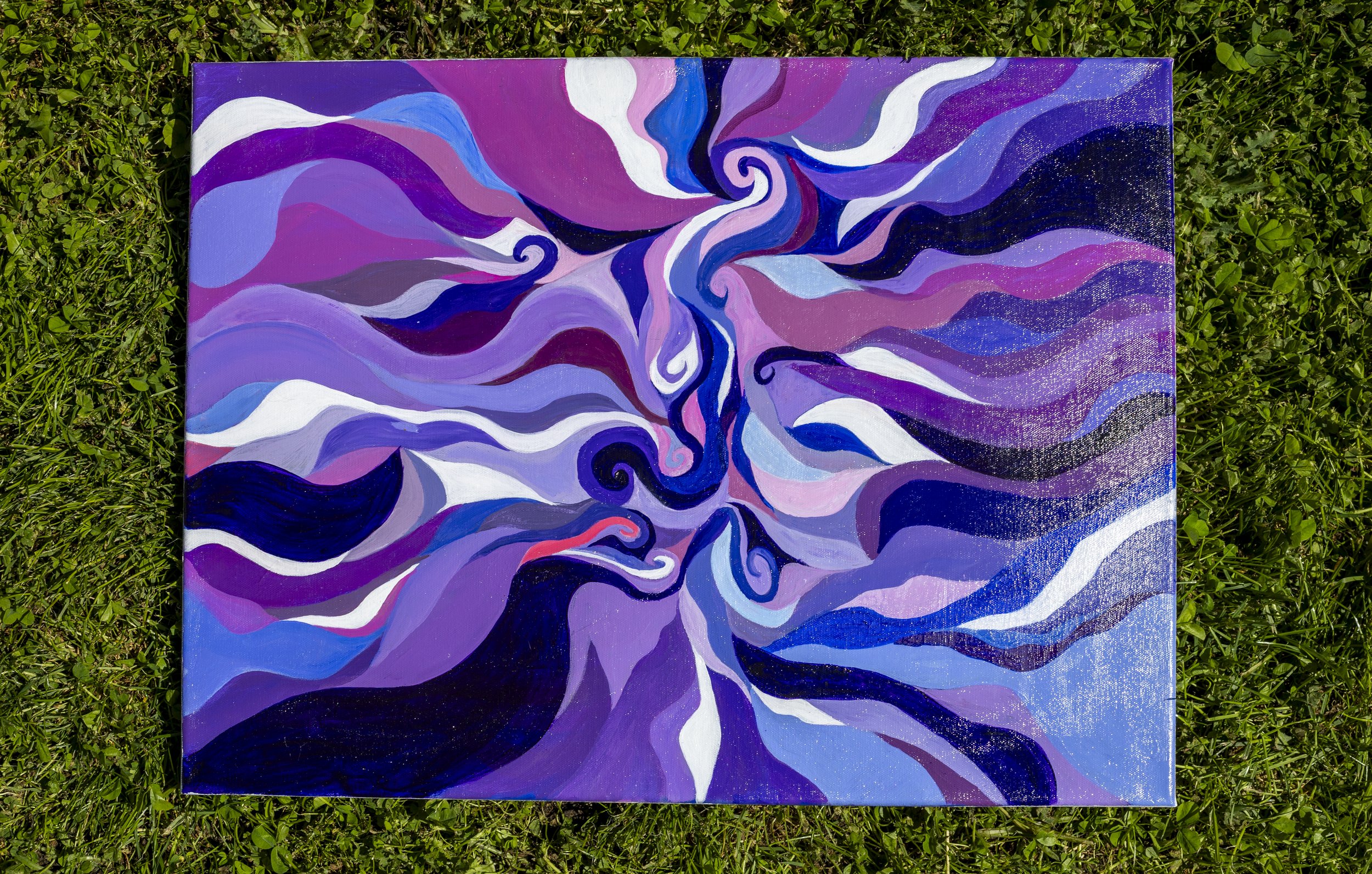An abstract acrylic painting with swirling patterns in shades of purple, blue, pink, white, and black, lying on green grass.