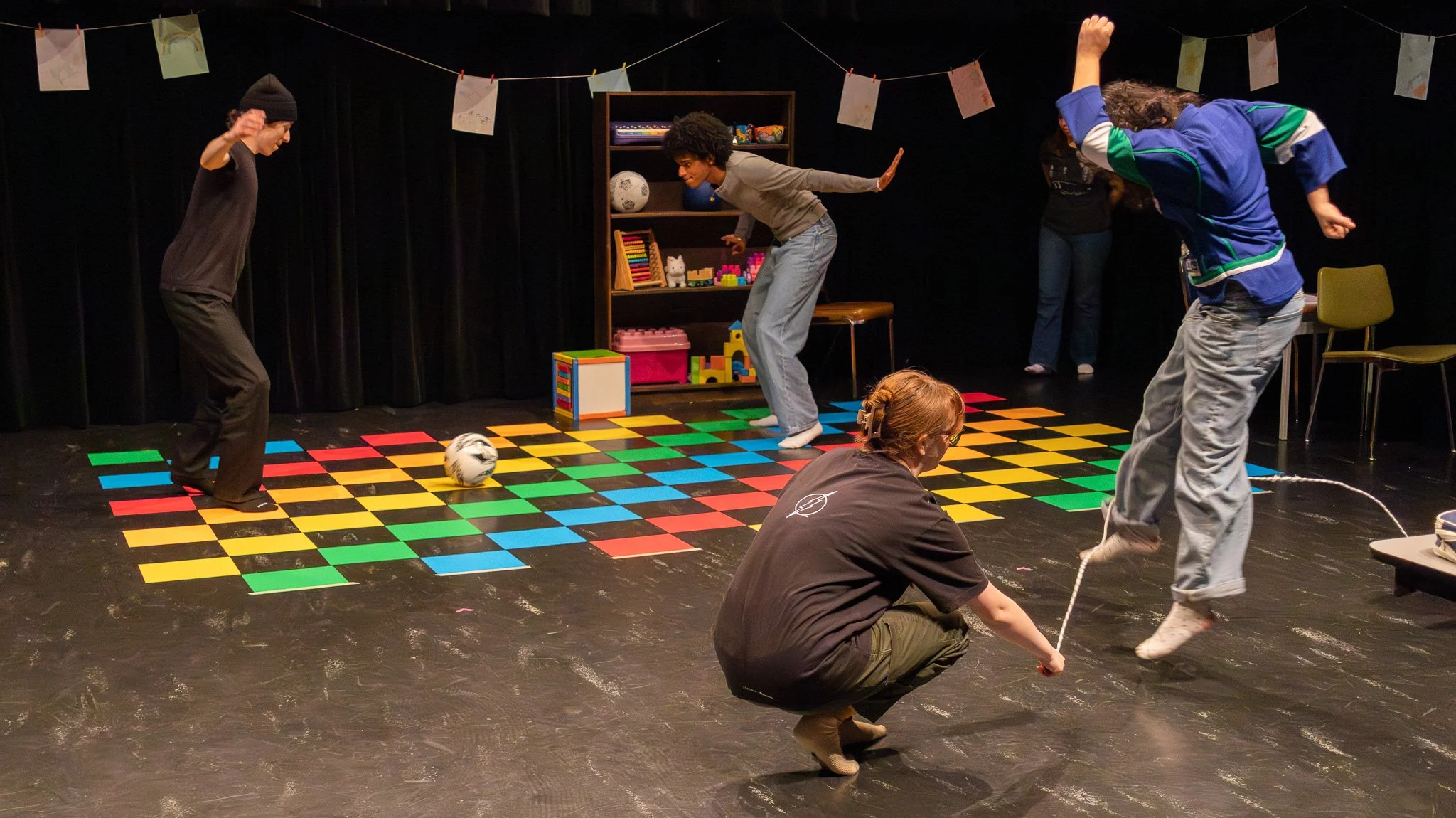 People dancing and playing on a colorful black and yellow checkered dance floor in a room with black curtains, shelves with toys, and children's artwork hanging overhead.