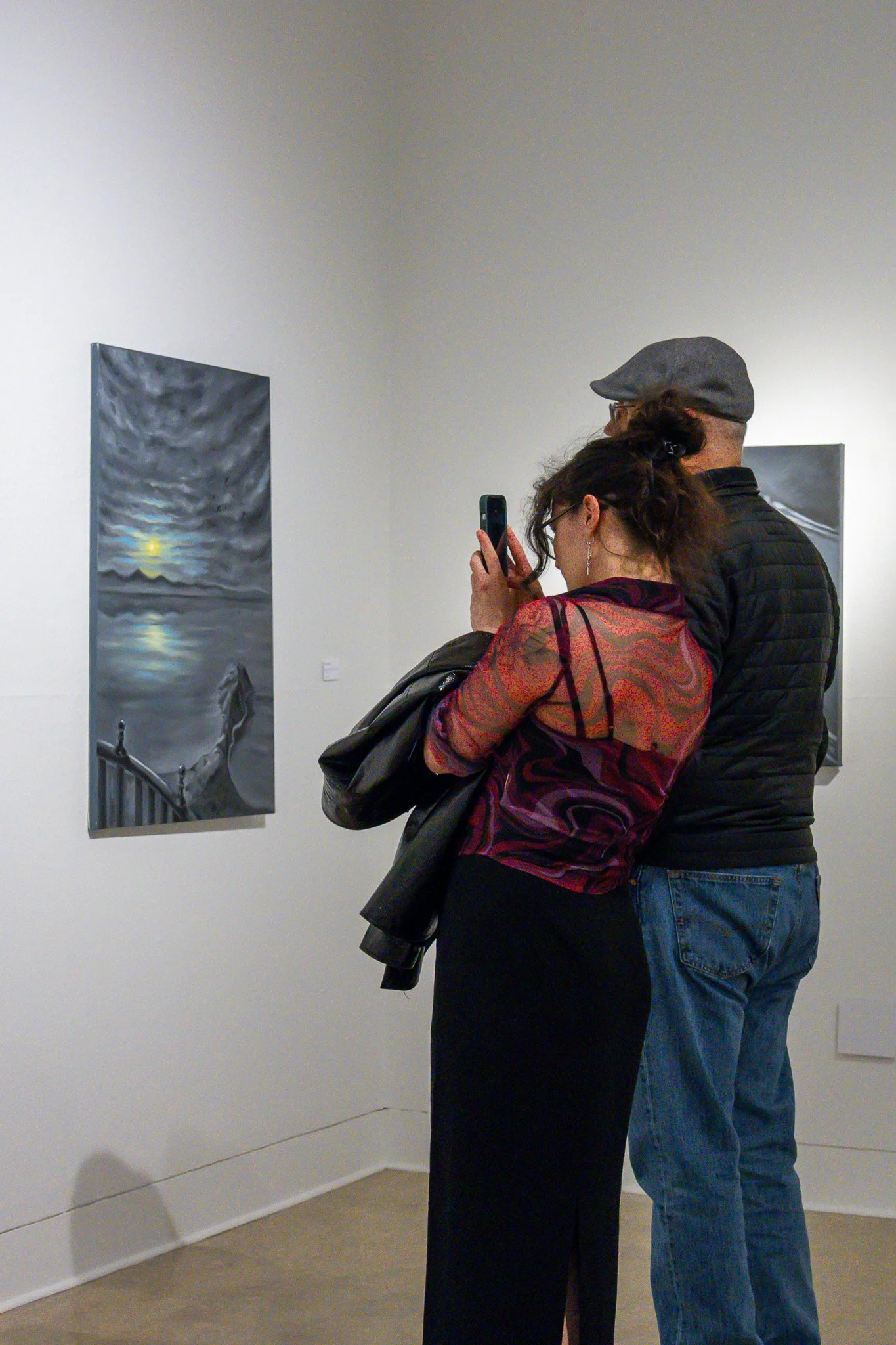 Two people standing in an art gallery, looking at artwork on the wall. The person on the left is a woman with dark hair, wearing glasses and a colorful patterned top, holding a phone. The person on the right is a man with a gray cap and glasses, wear