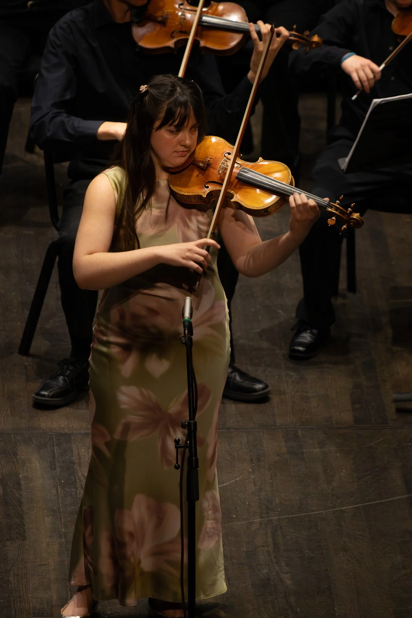 A woman in a floral dress playing the violin with an orchestra in the background.