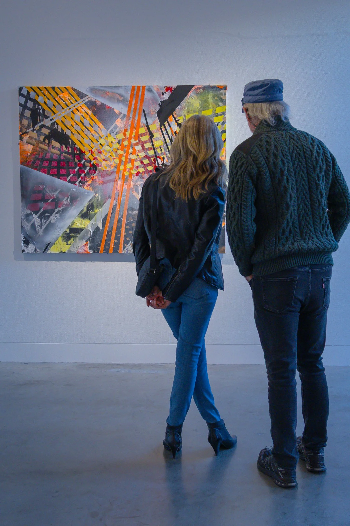 Two people, a woman and a man, are standing in front of a colorful abstract painting in a gallery. The woman has wavy blonde hair and is wearing a black leather jacket and blue jeans, while the man has long gray hair, glasses, a blue cap, and a dark 