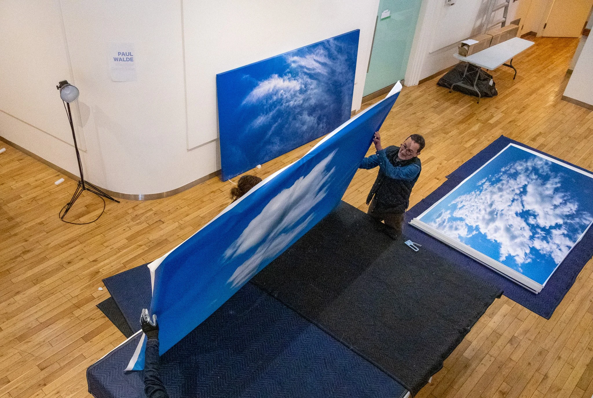 A person hanging a large canvas of a blue sky with white clouds on a wall in an art gallery or museum. There are additional canvases with similar sky images on the floor and a person sitting on a bench.