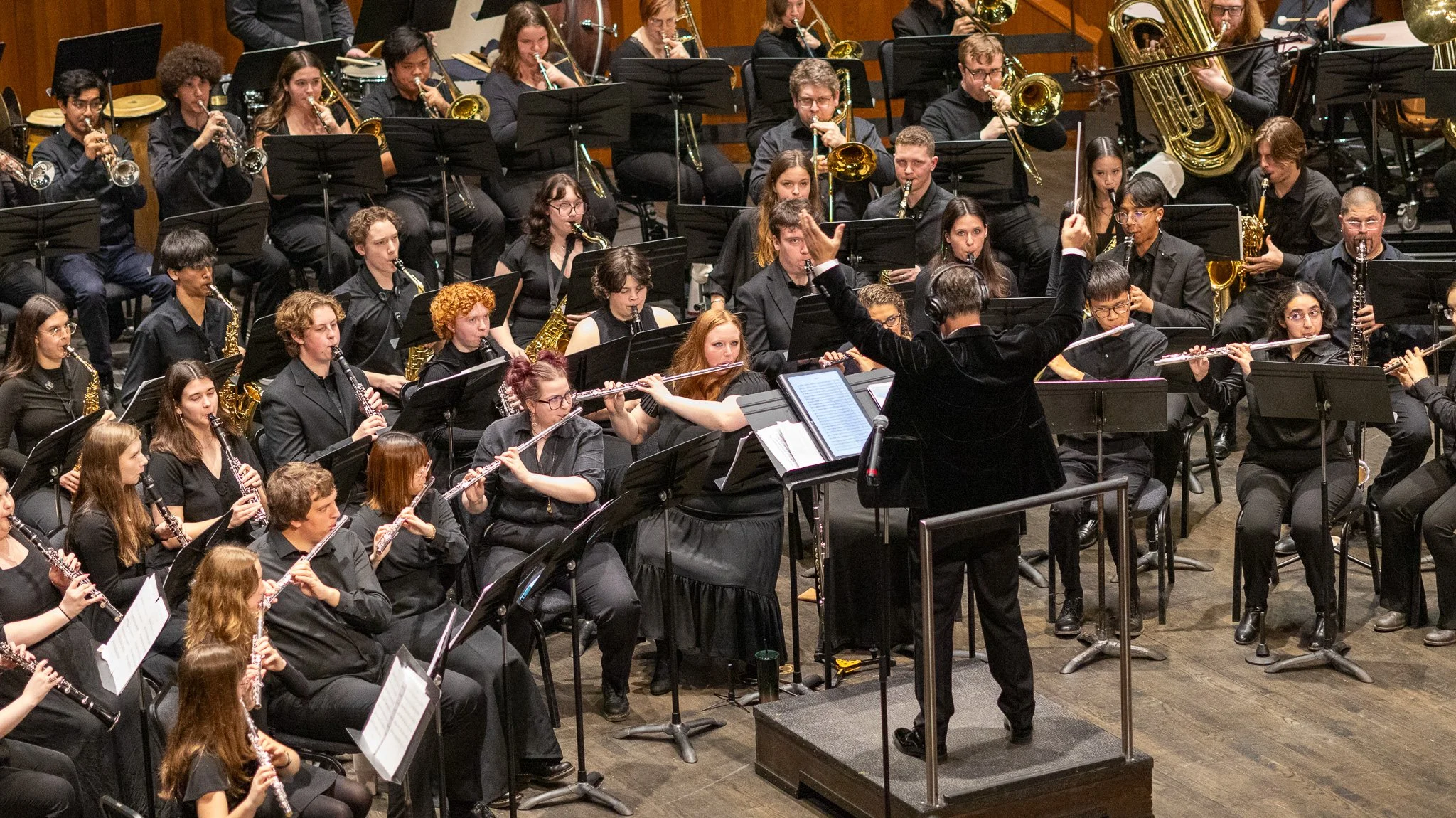 Orchestra performing on stage with musicians playing wind instruments, a conductor leading, and music stands with sheet music.