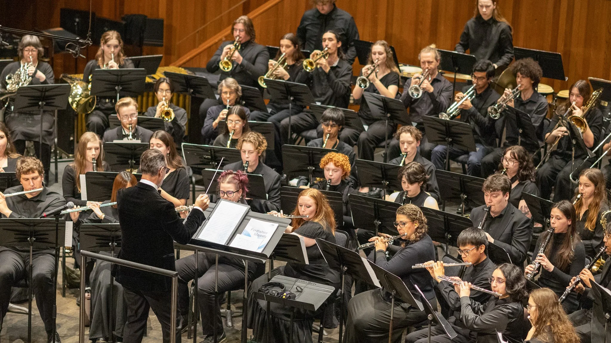 Orchestra performing on stage with musicians playing various instruments like trumpets, tubas, and flutes, conducted by a conductor in front.