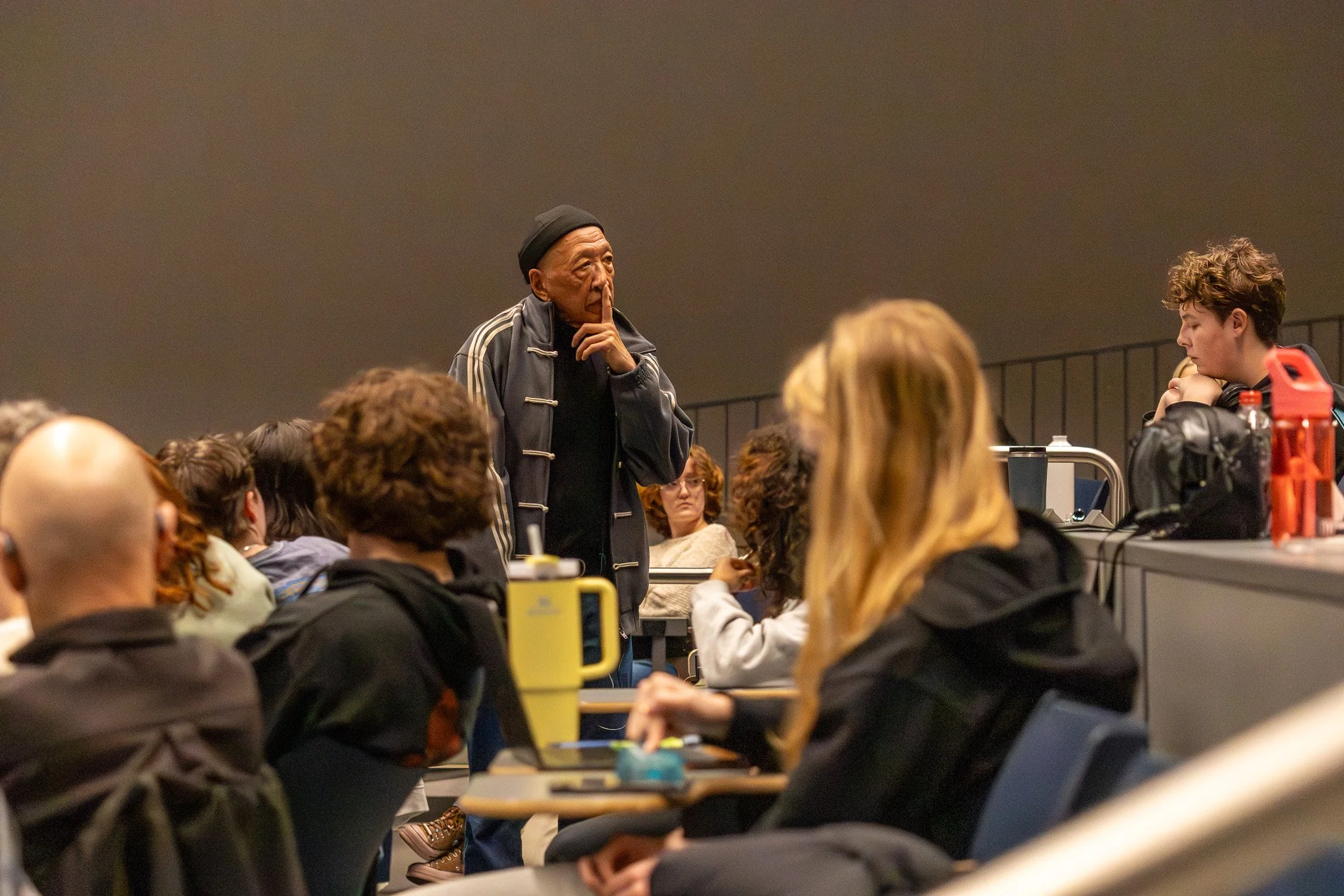 An elderly man with a finger near his lips stands in front of a seated group at a lecture or discussion, with some people taking notes or listening attentively.