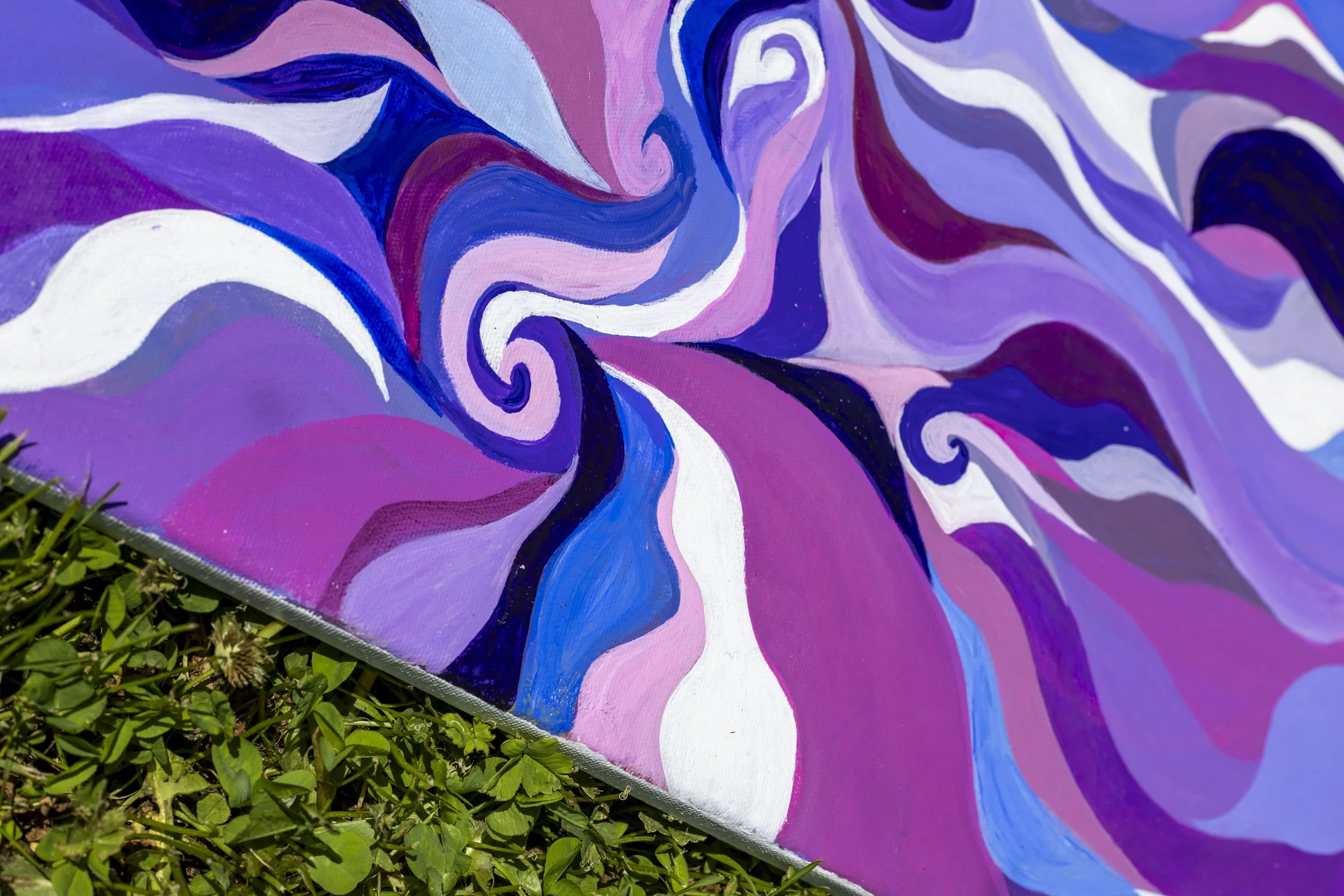 Close-up of an abstract, colorful painted mural featuring swirling patterns in shades of purple, pink, white, and blue, laid on grass.