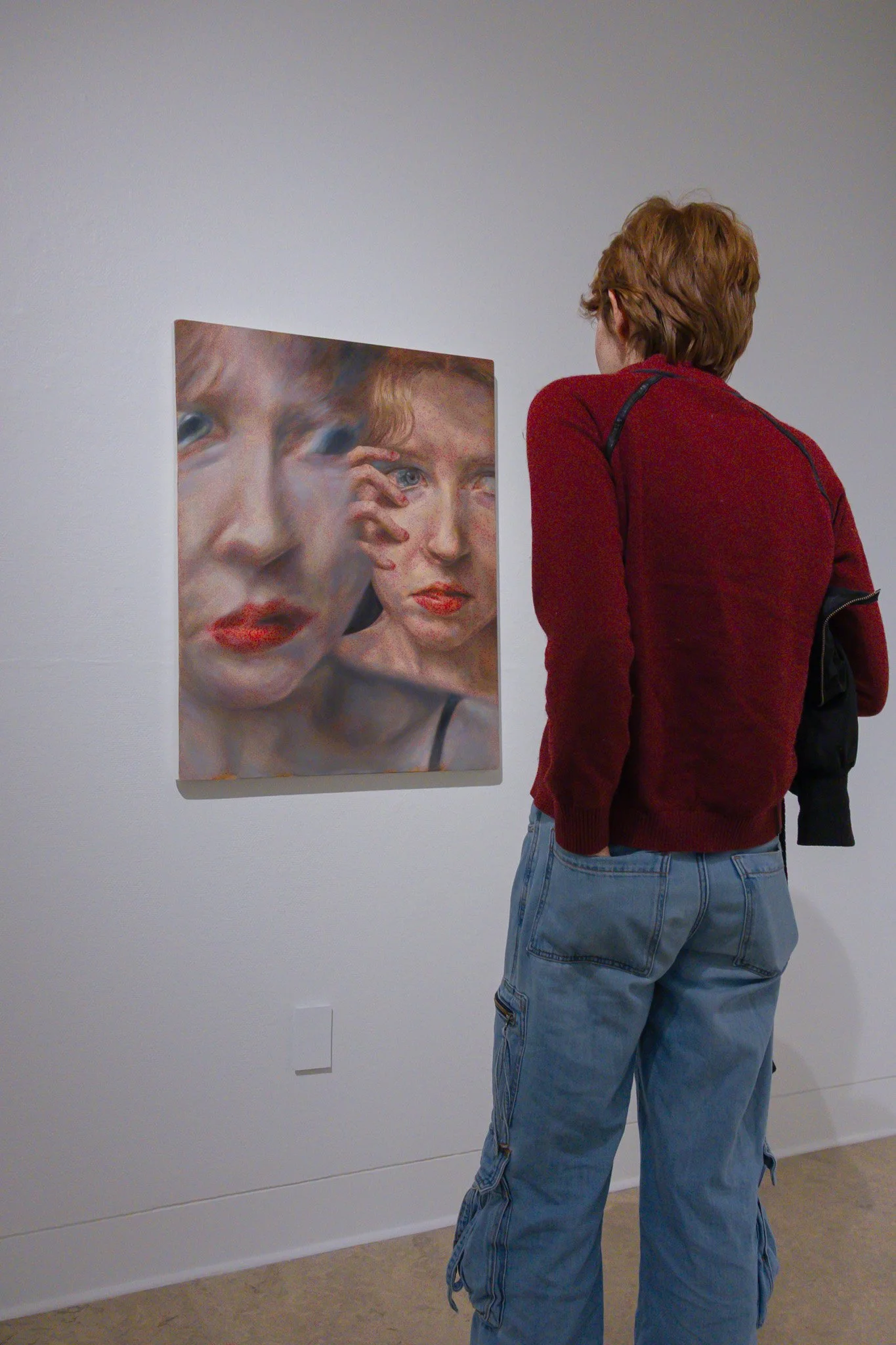 A person with short red hair wearing a red jacket and baggy jeans is looking at a large painting of two women's faces in an art gallery.