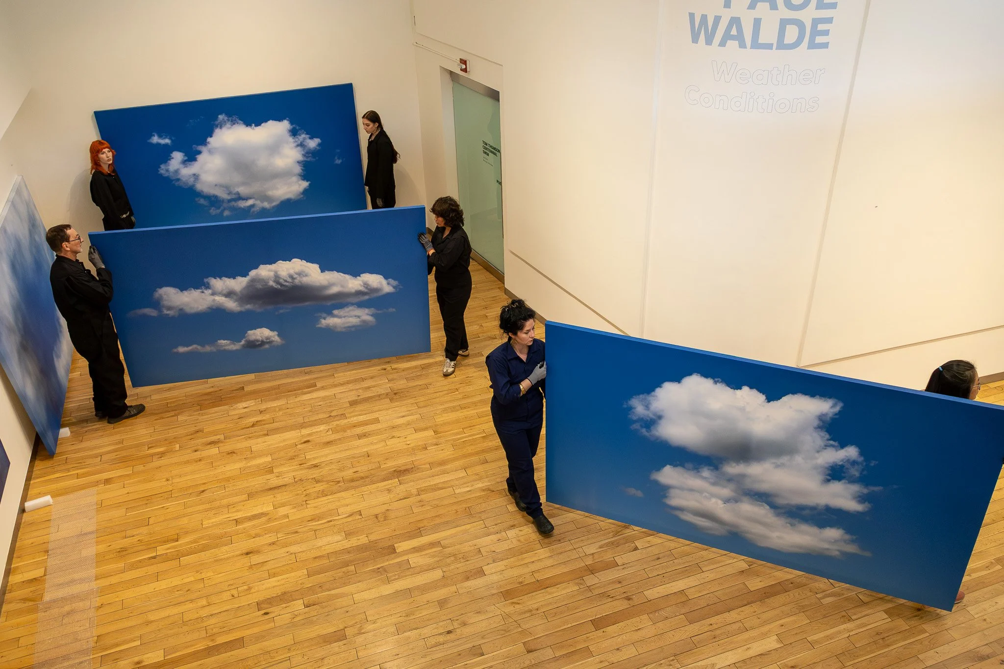 Several people are hanging large prints of blue skies with white clouds on a white wall in an art gallery.