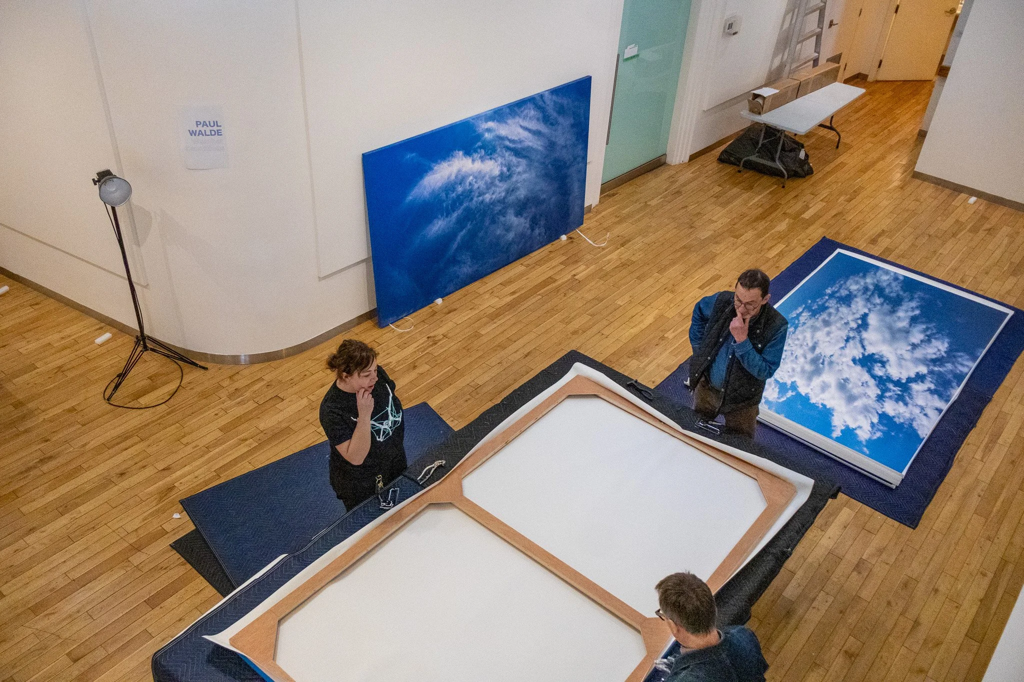 People preparing a photography setup with large prints of sky clouds, one leaning against a wall and others standing around a table in a room with wooden floors.