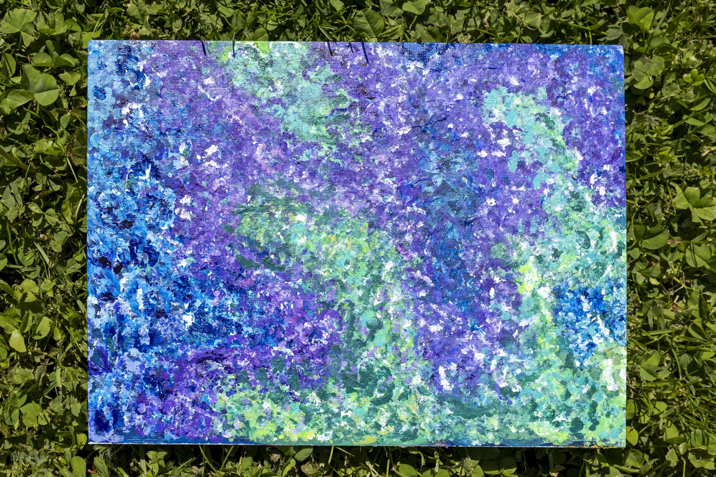 Close-up of a textured, abstract painting with shades of blue, purple, green, and white, placed on green foliage.