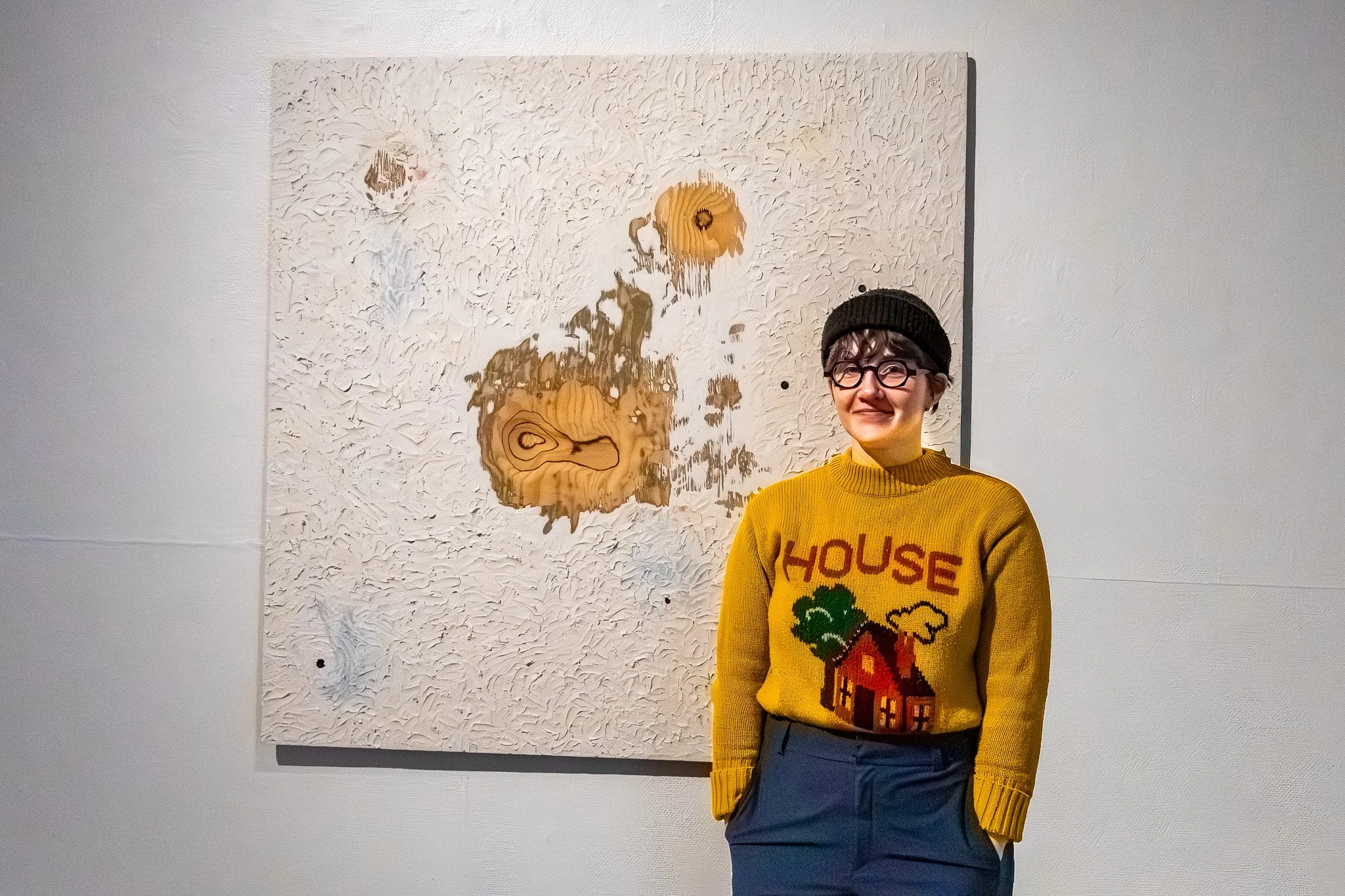 Young woman with short hair, glasses, and a black beanie, wearing a yellow sweater with a graphic and the words 'HOUSE MUSIC,' standing in front of an abstract textured painting on a white wall.