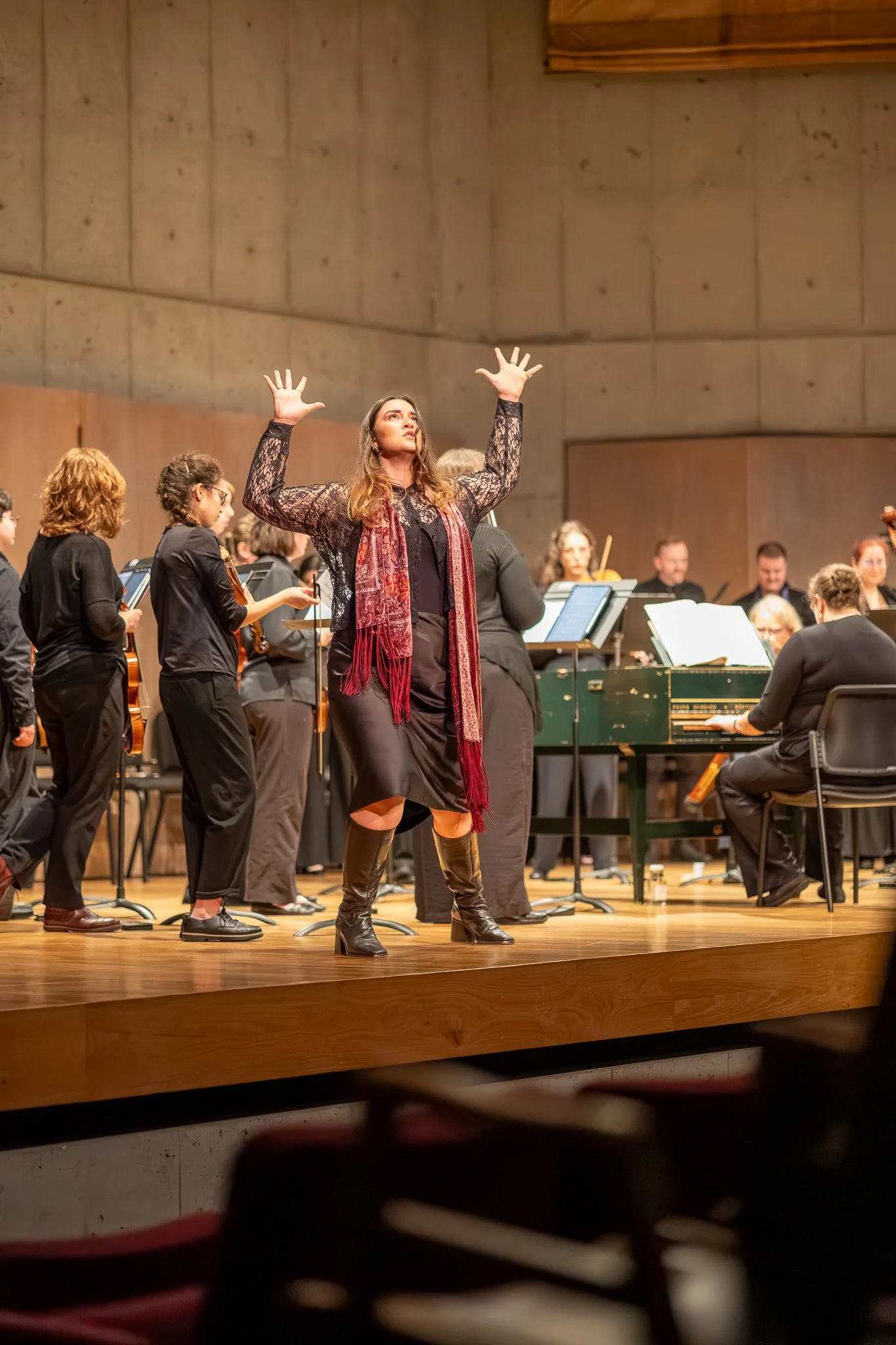 A woman standing on stage with her arms raised, leading an orchestra in a concert hall. The orchestra players are intently focused on their music, with some holding their instruments. The woman appears confident and engaged in conducting.