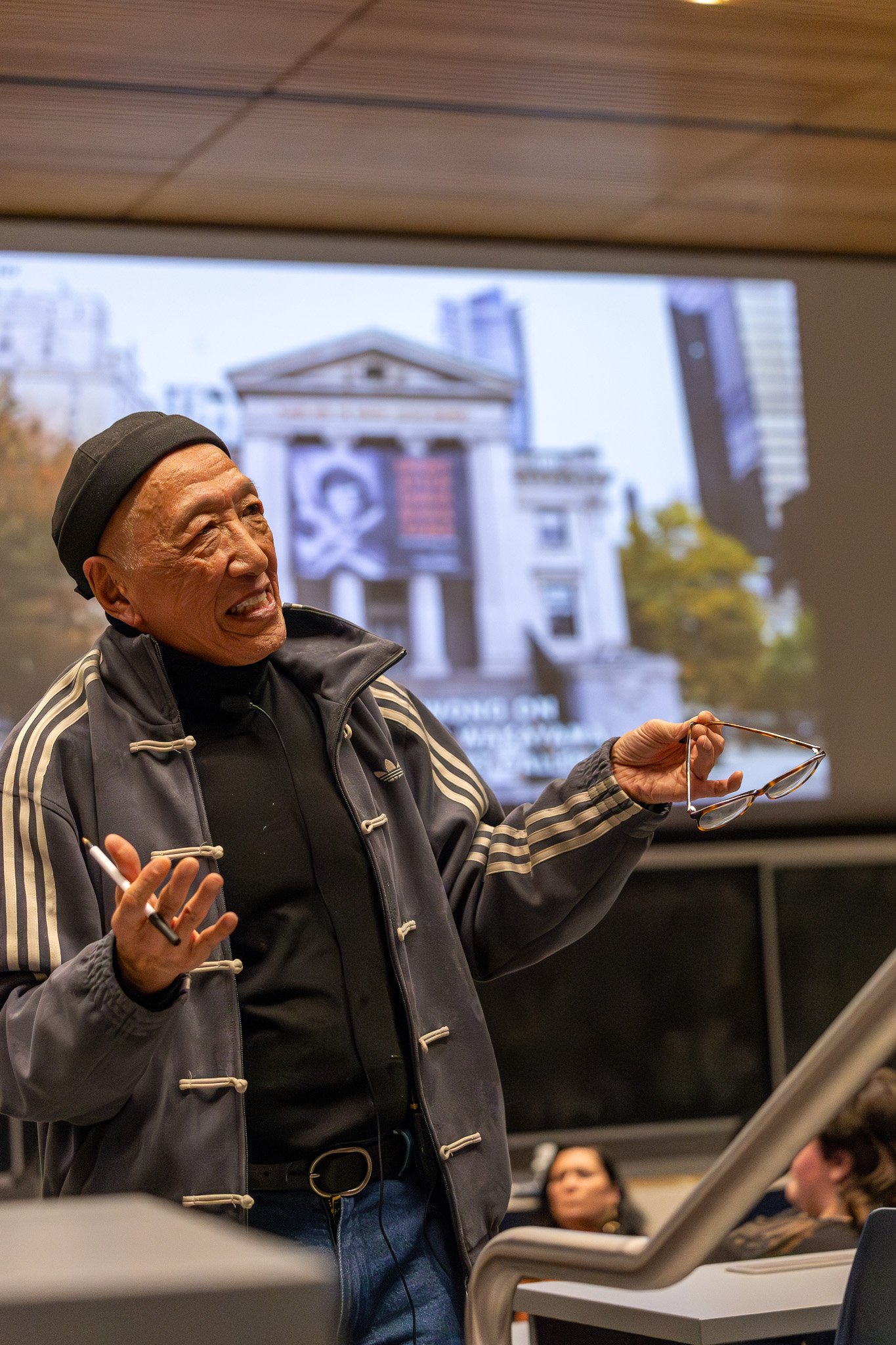 Elderly man laughing and speaking, holding reading glasses in hand, in a classroom or seminar setting with other attentive participants and a projected image of a building on the screen behind him.