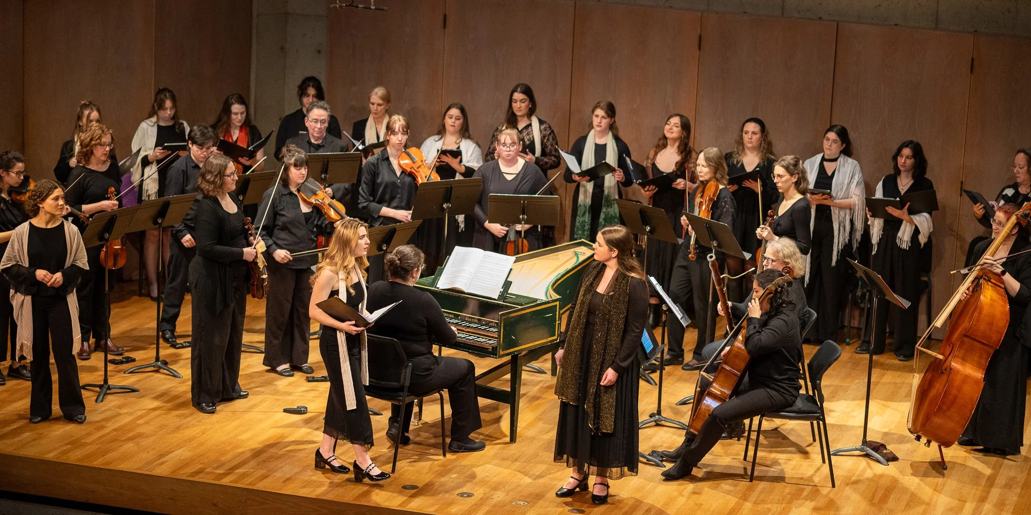 An orchestra performing on stage, with musicians playing violins, a cello, and a grand piano, under warm stage lighting.