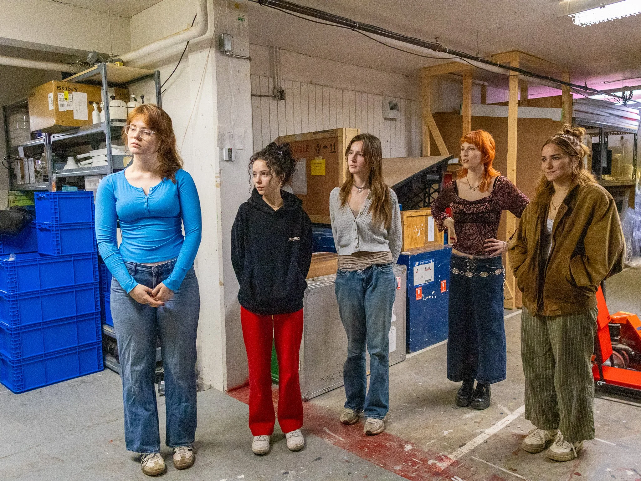Five young women standing in a warehouse or storage room, facing different directions. The background shows shelves with boxes and storage containers, and wooden frames.