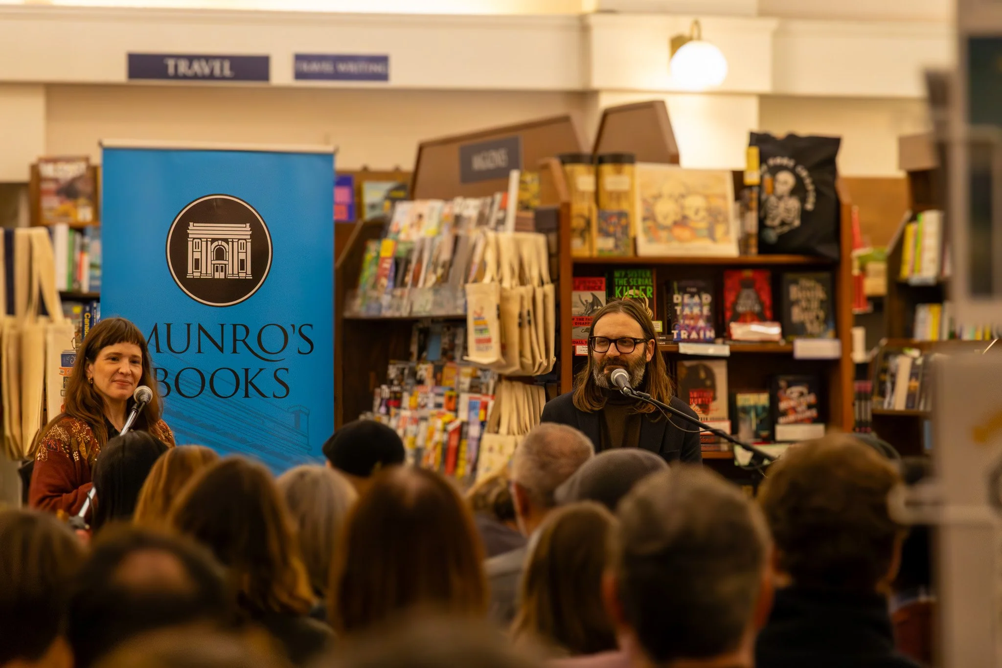 A man with long hair and glasses speaking into a microphone in a bookstore with shelves of books and a blue banner reading "Munro's Books." A woman stands nearby. Audience members are visible in the foreground.