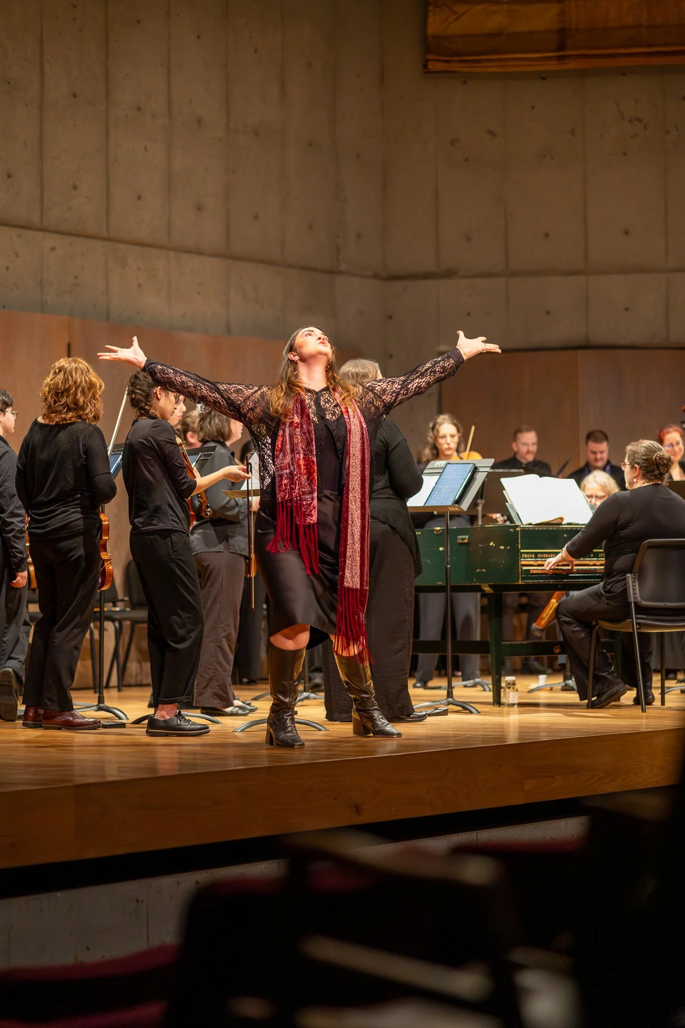A woman with long hair, wearing a black lace top and leather boots, stands with arms outstretched on a stage with an orchestra behind her. The orchestra consists of musicians playing string instruments, and a pianist on a green piano. The stage has a