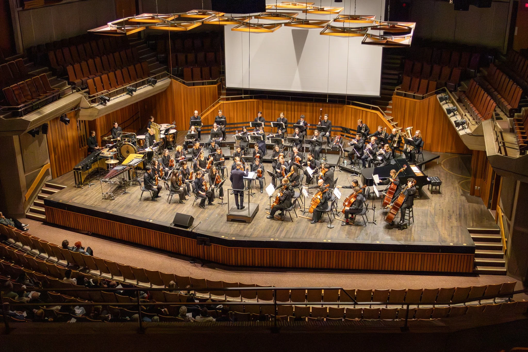 An orchestra performing on a stage in a concert hall with a conductor leading the musicians. The hall has wooden accents and audience seating visible in the foreground.