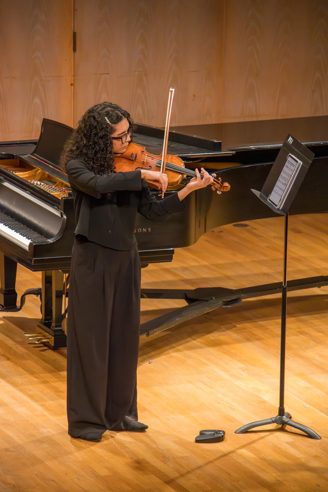 A woman playing the violin on stage with music sheet on a stand and a grand piano in the background.