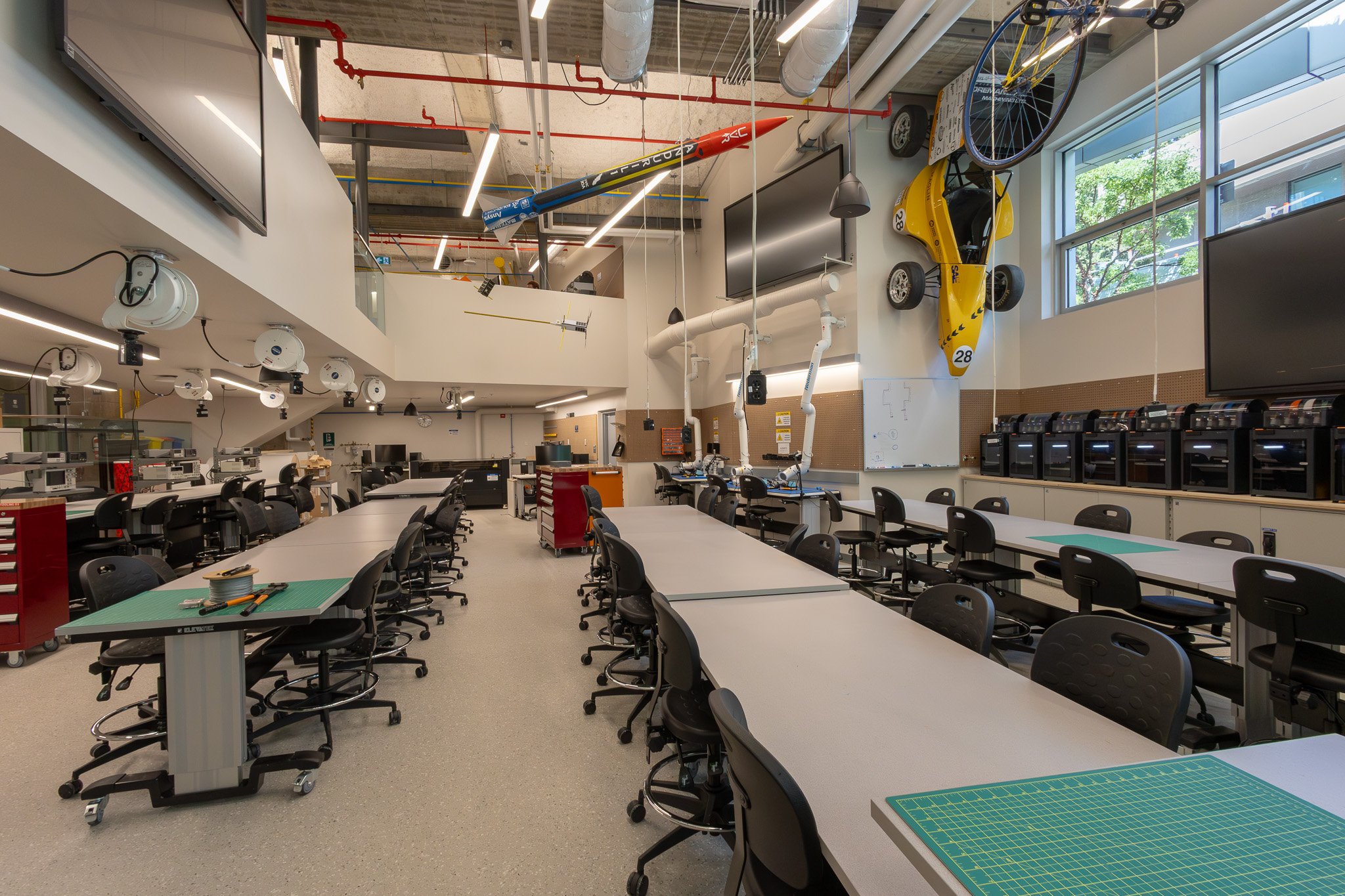 A spacious, modern classroom or lab with rows of tables and chairs, featuring projectors, multiple computers, and a large window with trees outside. The ceiling has industrial exposed ductwork and hanging lights, with model airplanes and a bicycle ha