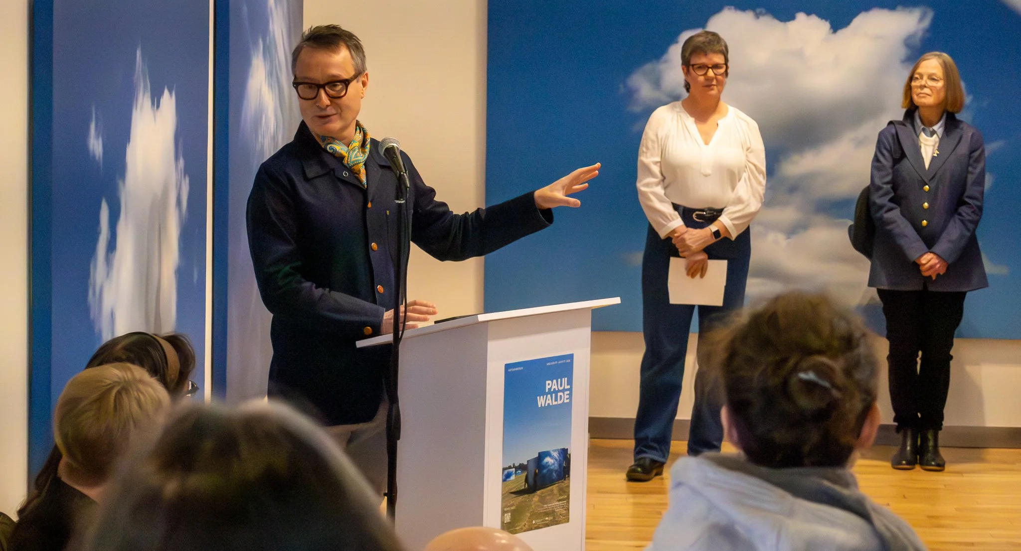 A man wearing glasses and a dark blazer is speaking at a podium, gesturing with his hand. Two women are standing in the background: one in a white blouse and the other in a blue blazer, both watching him. There are children in the foreground and a bl