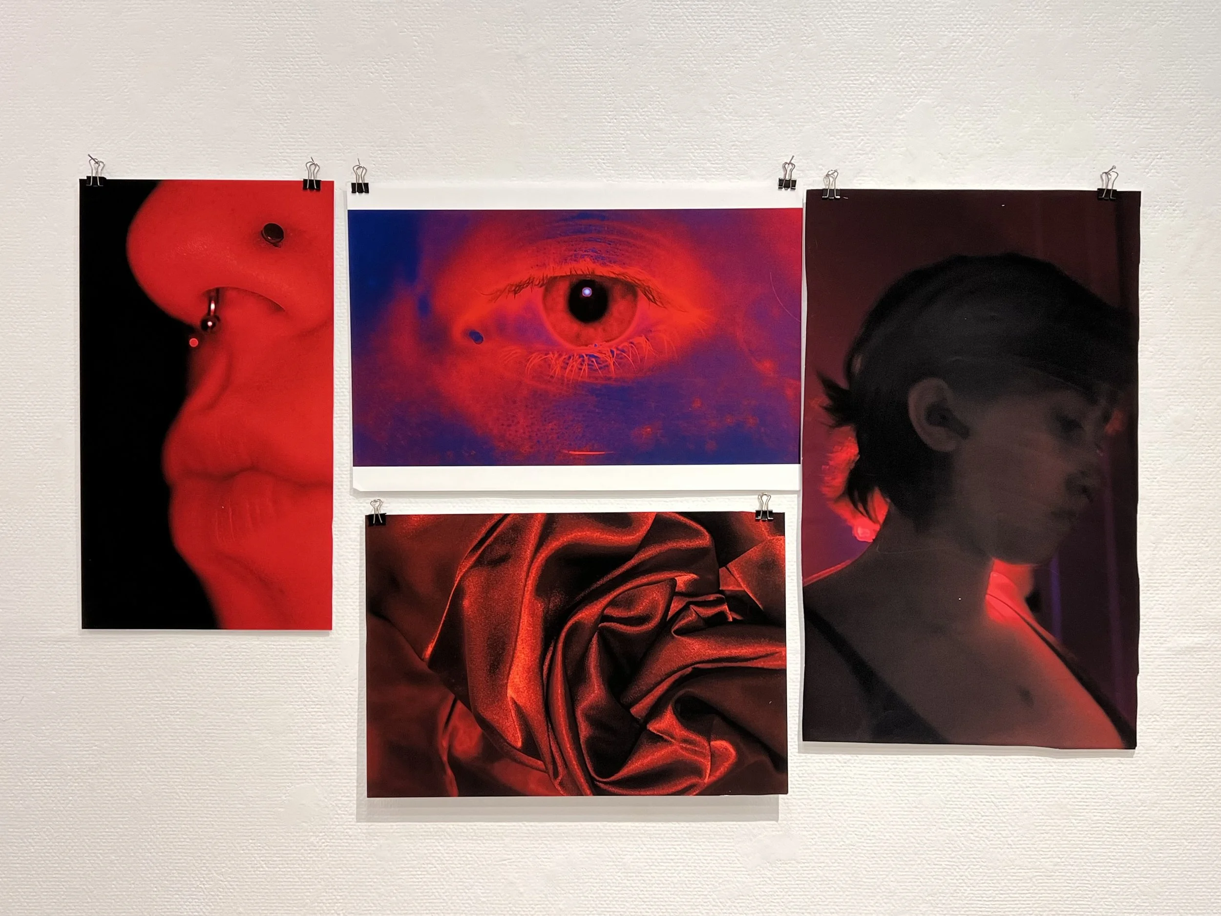 Three photographs displayed on a beige wall, featuring a red-tinted close-up of an eye in the center, a person with a shaved head and dreadlocks on the right, and a person with facial jewelry on the left.