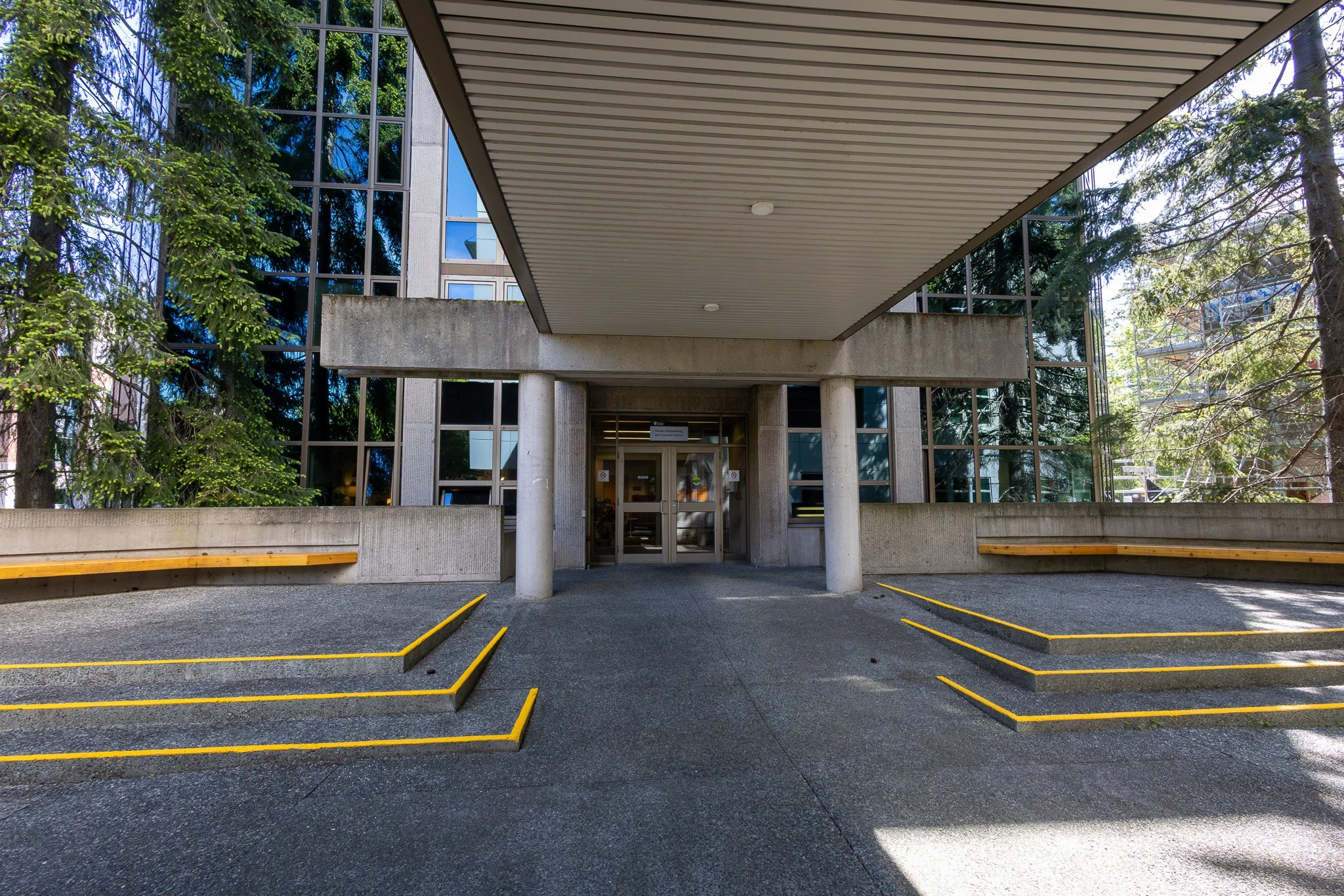 Entrance to a concrete building with large glass windows, surrounded by trees, with yellow-lined parking spaces in the foreground.