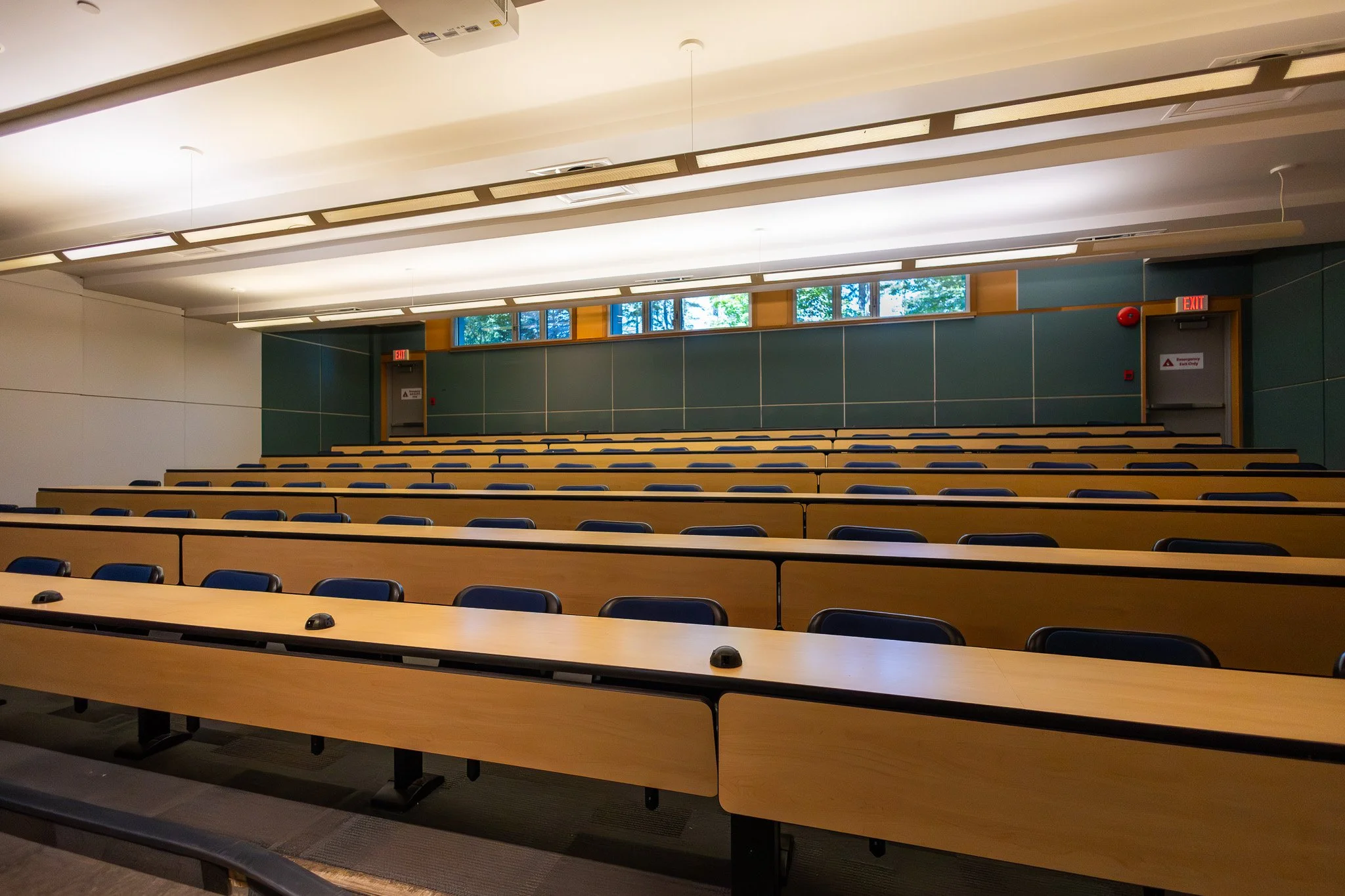 Empty classroom or lecture hall with long rows of wooden desks and blue chairs, large windows with trees outside, ceiling with panels and lighting, and doors with exit signs.