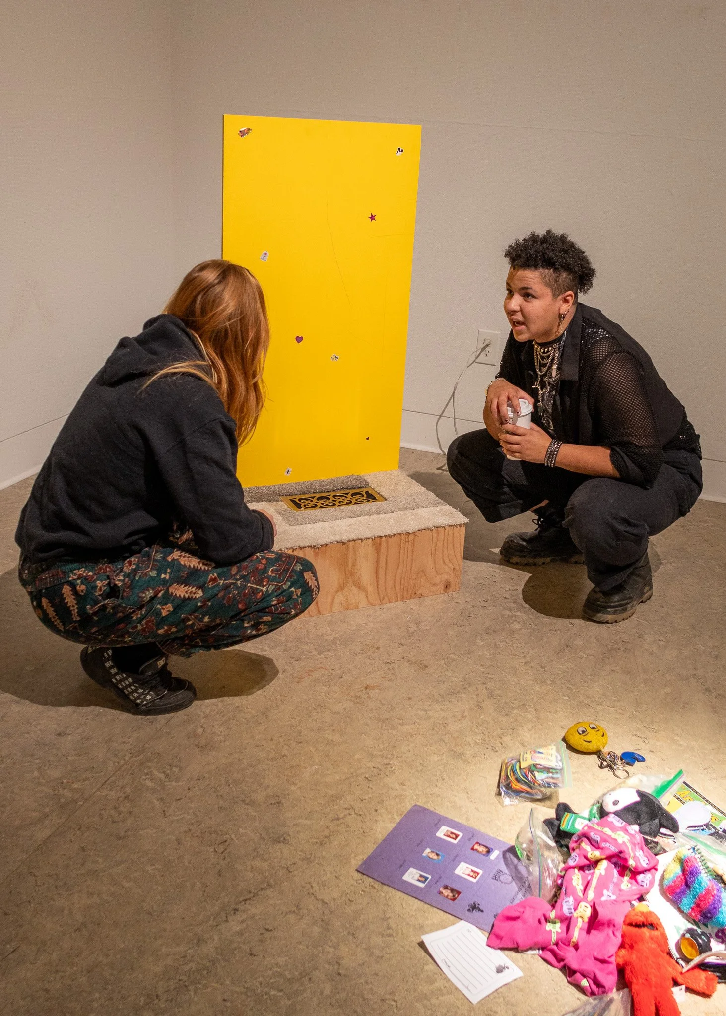 Two women engaging in conversation in front of a yellow wall. One is kneeling on the floor, the other is squatting, and there are various items on the floor including clothes, toys, and papers.