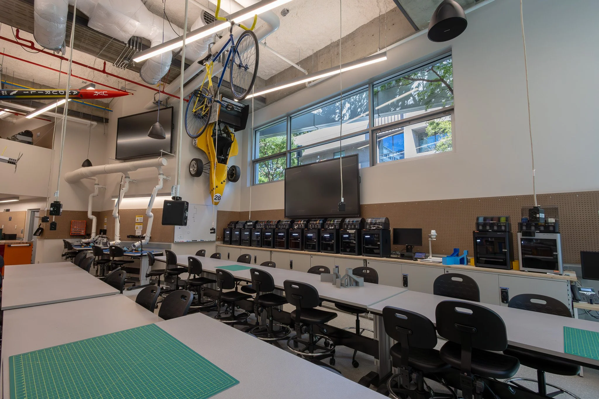 A modern classroom or workshop with tables and chairs, computer stations, a large window, and various equipment including a yellow bike, a yellow go-kart, and a red rocket model hanging from the ceiling, with fluorescent lighting and industrial-style