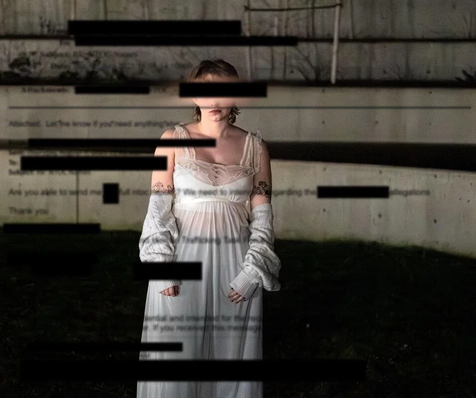 A woman in a white dress with tattoos on her arms stands outdoors near a wall with black bars and blurred text, under dim lighting.
