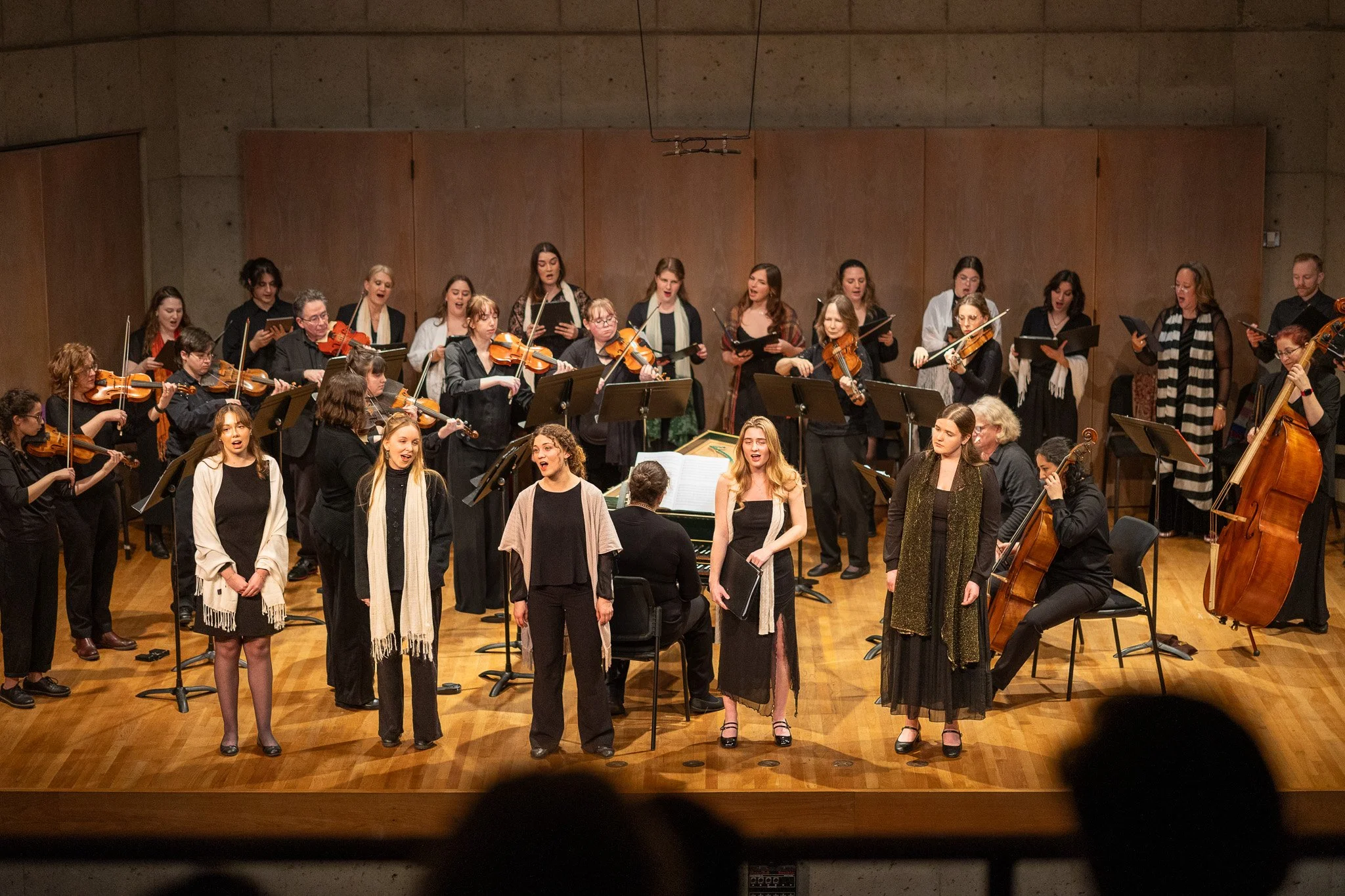 A group of musicians and singers performing on stage, with some playing string instruments like violins and a double bass, and others singing, in a concert hall with a wooden floor and a plain wall background.