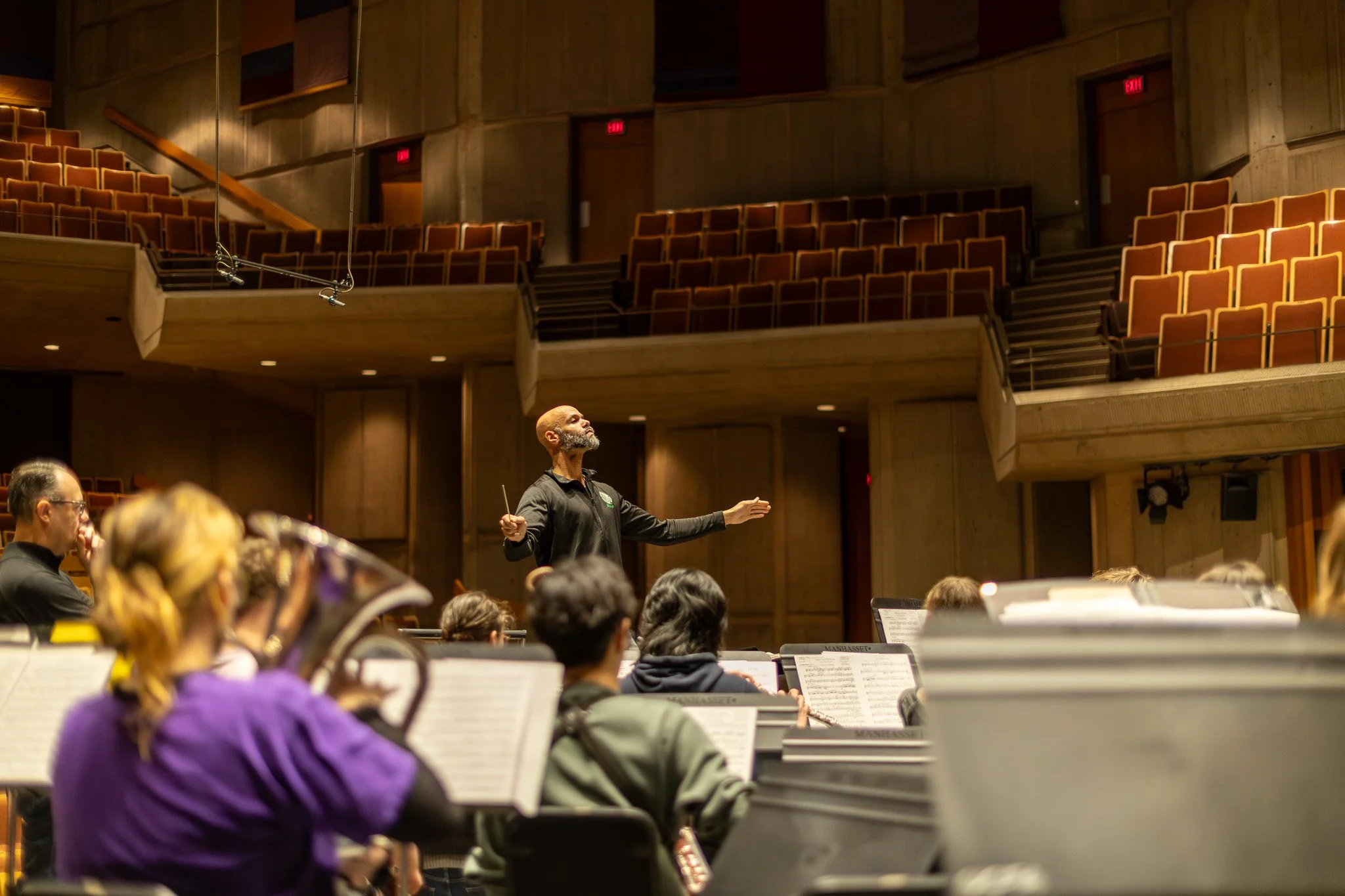 A conductor leading an orchestra in a concert hall with tiered seating and wood-paneled walls.