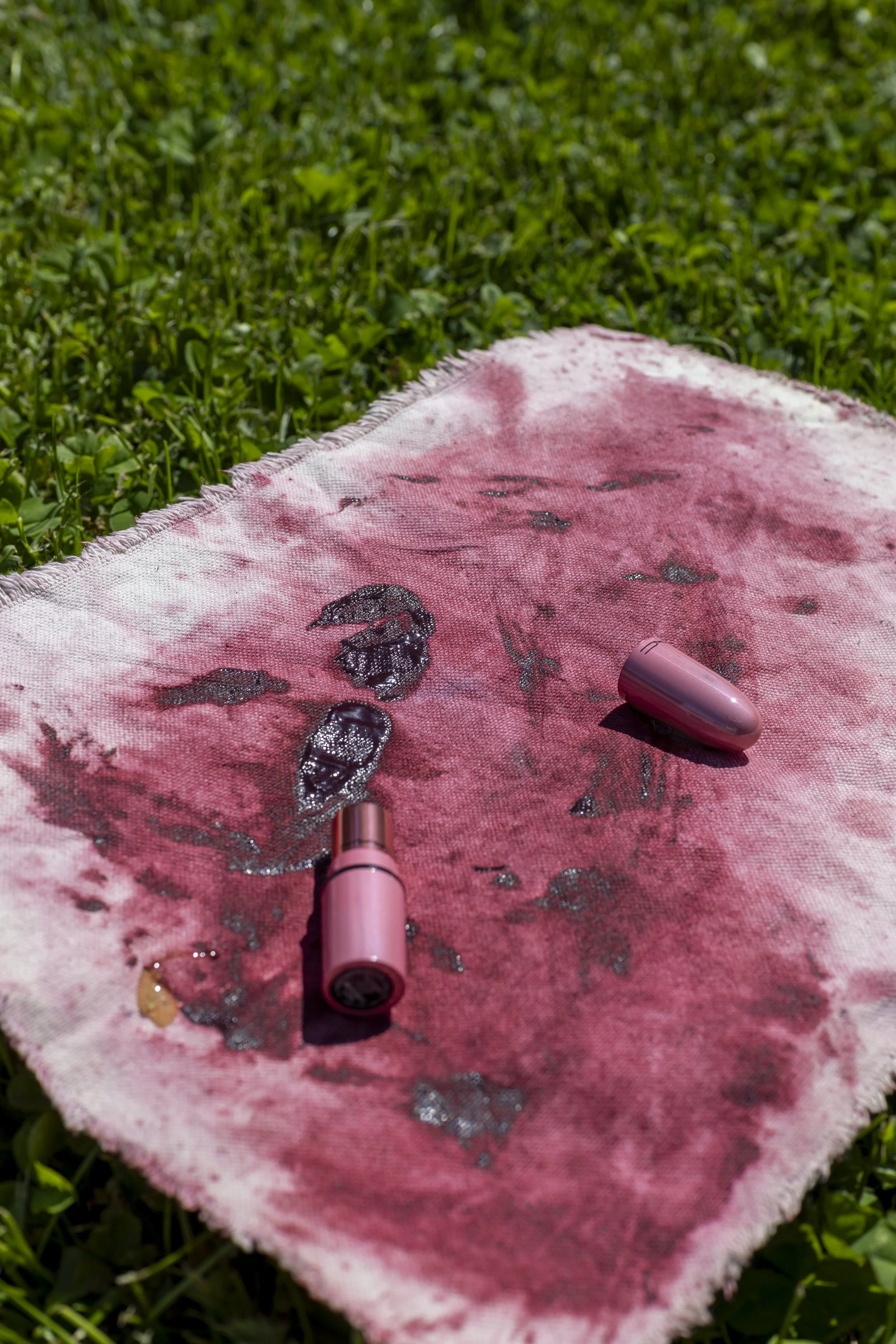 Pink towel with black liquid stains, a pink vape, and a pink lipstick, on green grass.