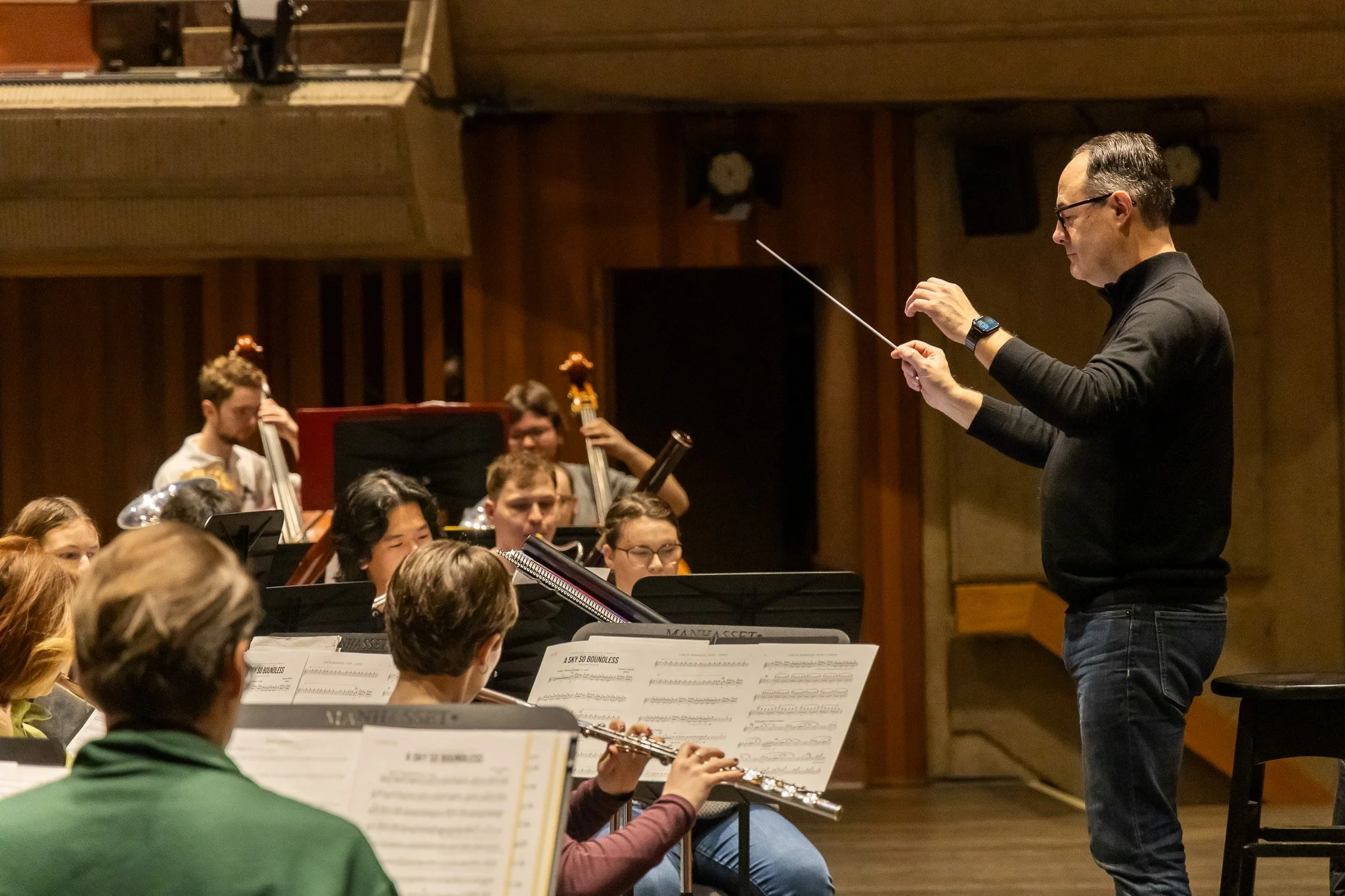 A conductor leads an orchestra during a rehearsal, with musicians reading sheet music and various instruments present in a wooden-paneled concert hall.
