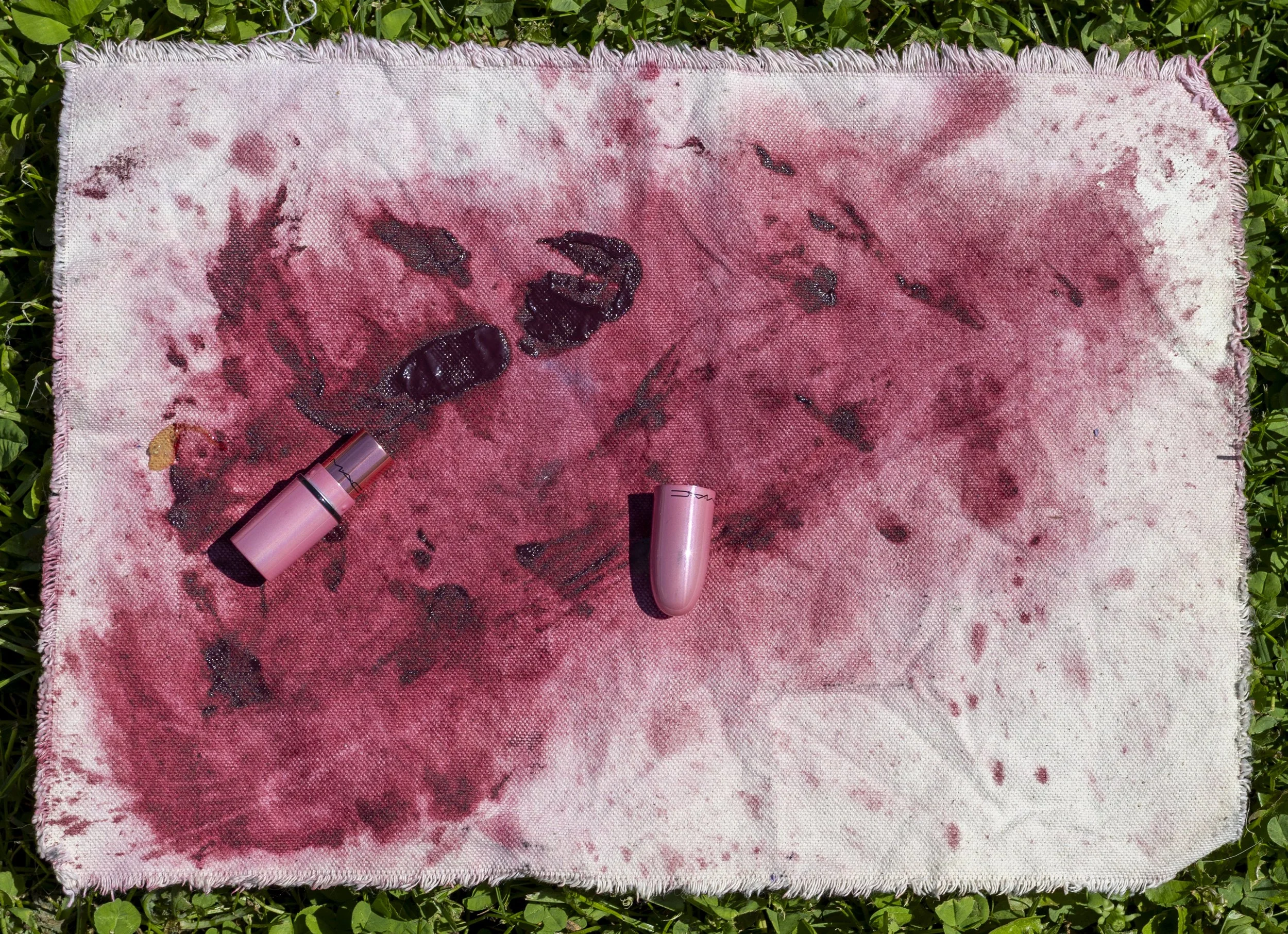 A blood-stained towel on the grass has several blacked-out fragments, a black pill, and some lipsticks.