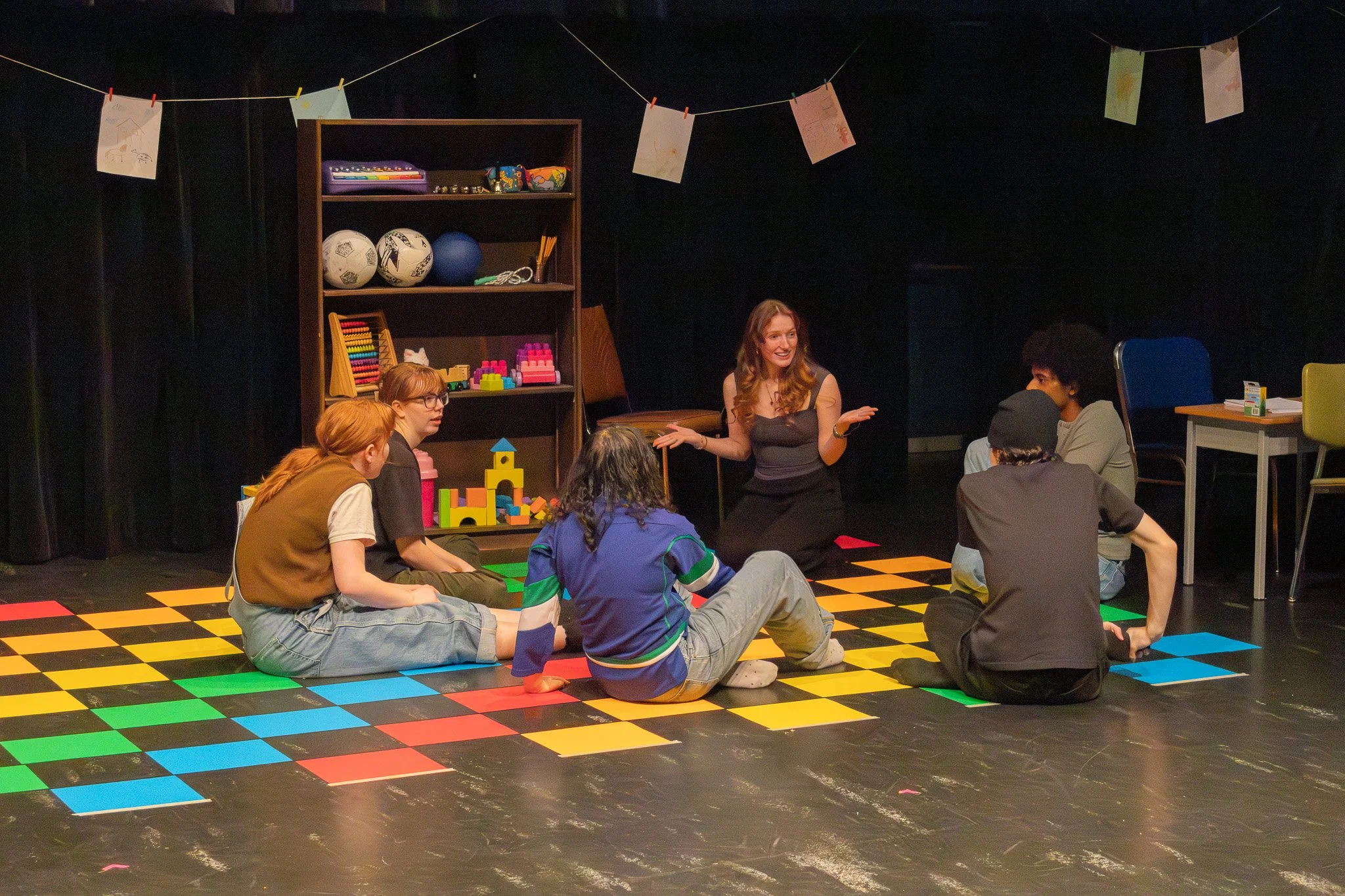 A woman with long hair, sitting and talking to a group of young people gathered on a colorful checkered floor in a dark room, with a black background and a bookshelf with soccer balls and toys behind her, while colorful paper coats are hanging from a