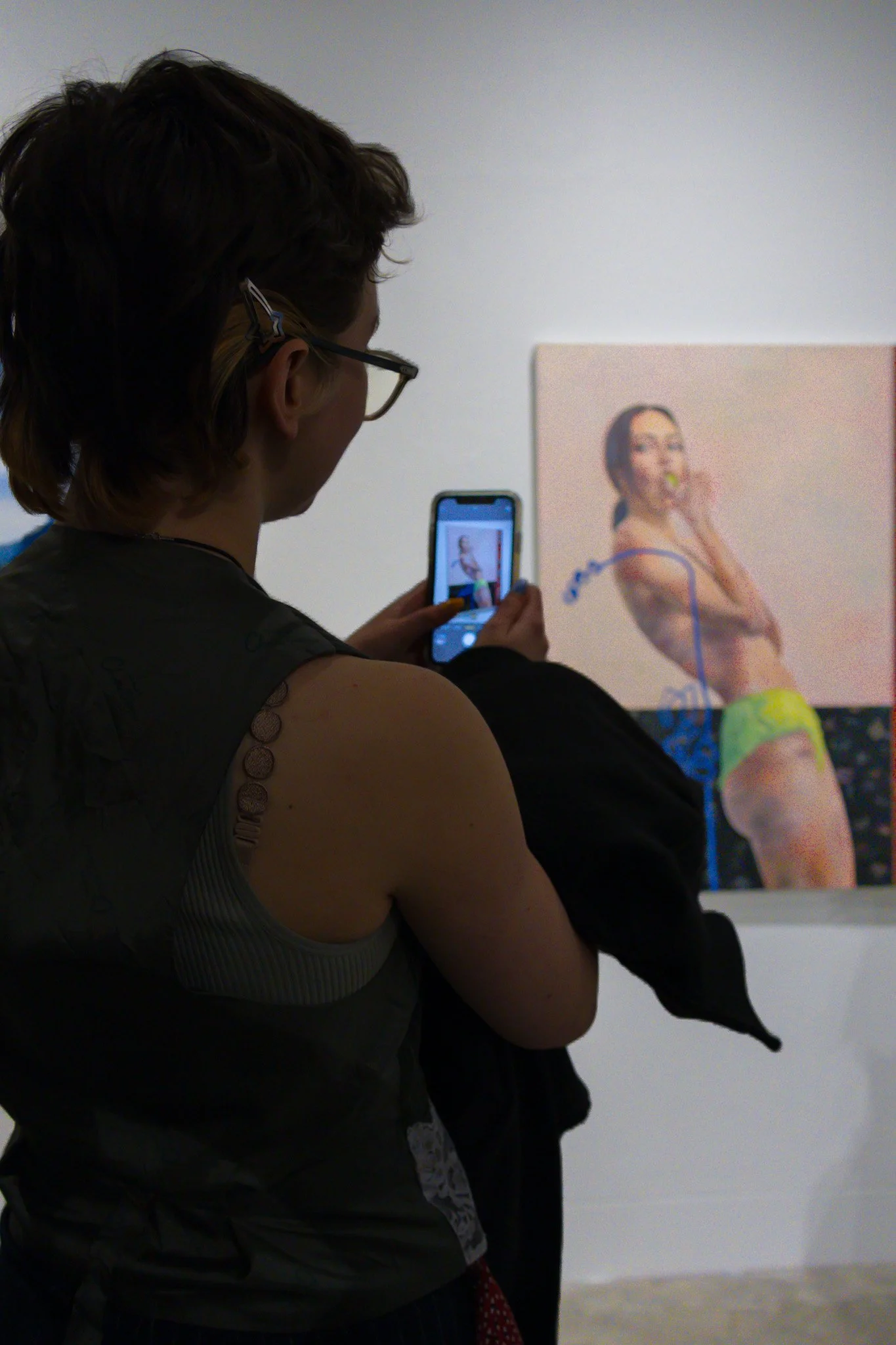 A woman with glasses and short hair taking a photo of a painting of a woman with long dark hair, holding a tennis ball near her mouth, in an art gallery.