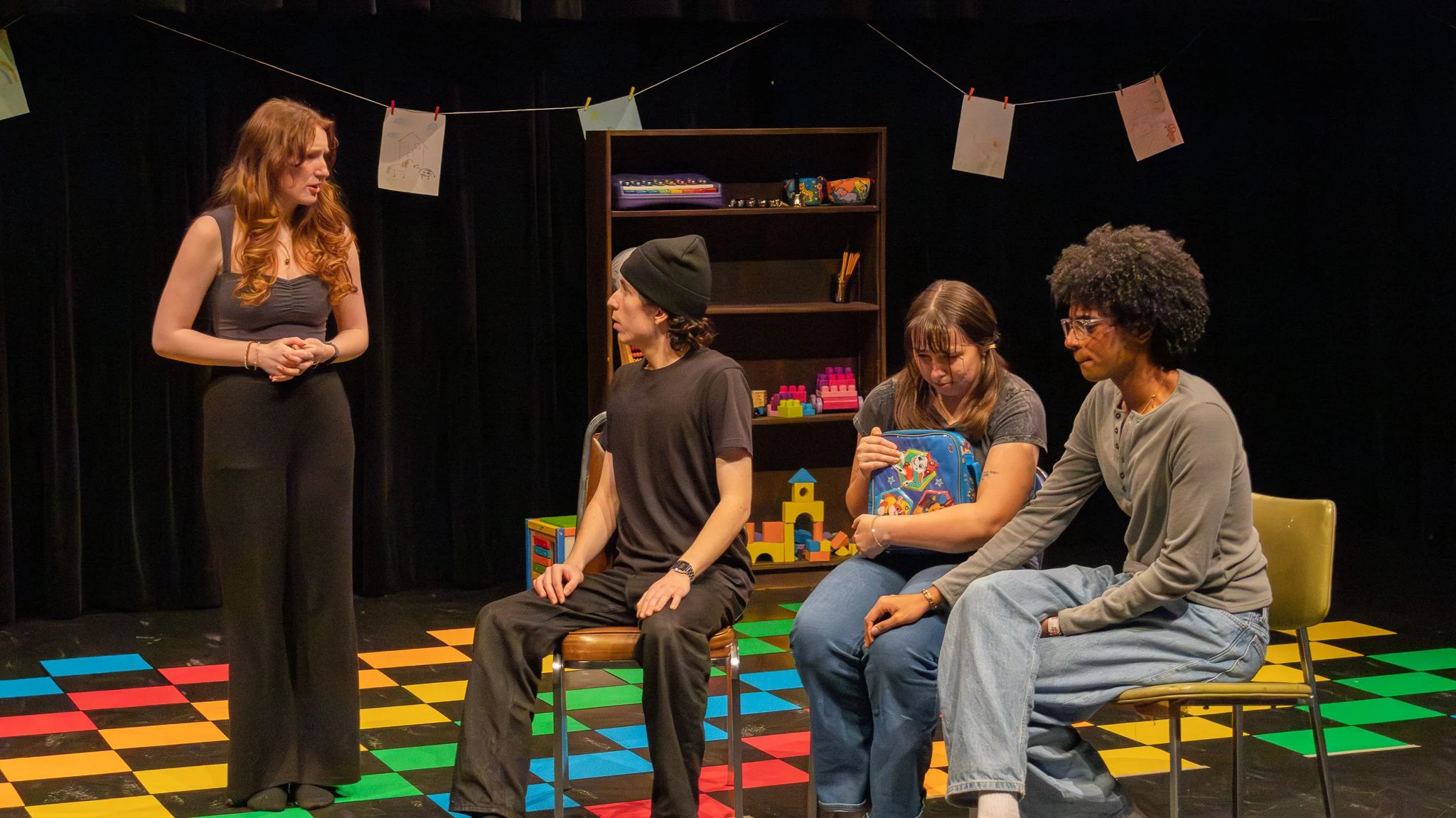 Four people seated in a room with a dark curtain background and colorful square floor pattern. A woman with long red hair stands and talks to the three seated individuals, who appear focused on her. There is a shelf with toys and drawings hanging on 