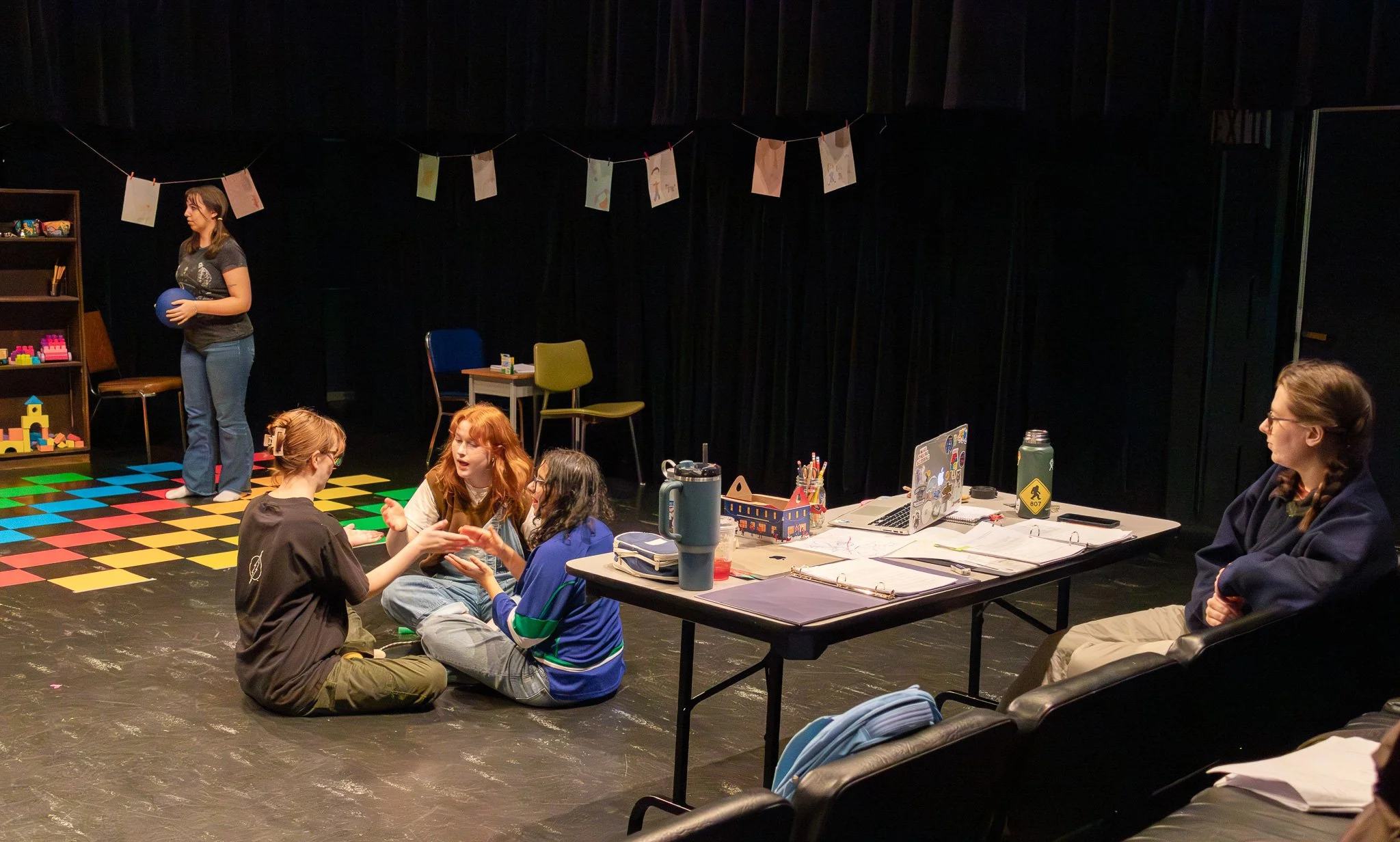 A group of young people and a woman with long hair and glasses sitting and chatting at a table with laptops and papers on it. In the background, a young woman standing on a colorful checkered floor holding a blue ball, with arts and crafts hanging on
