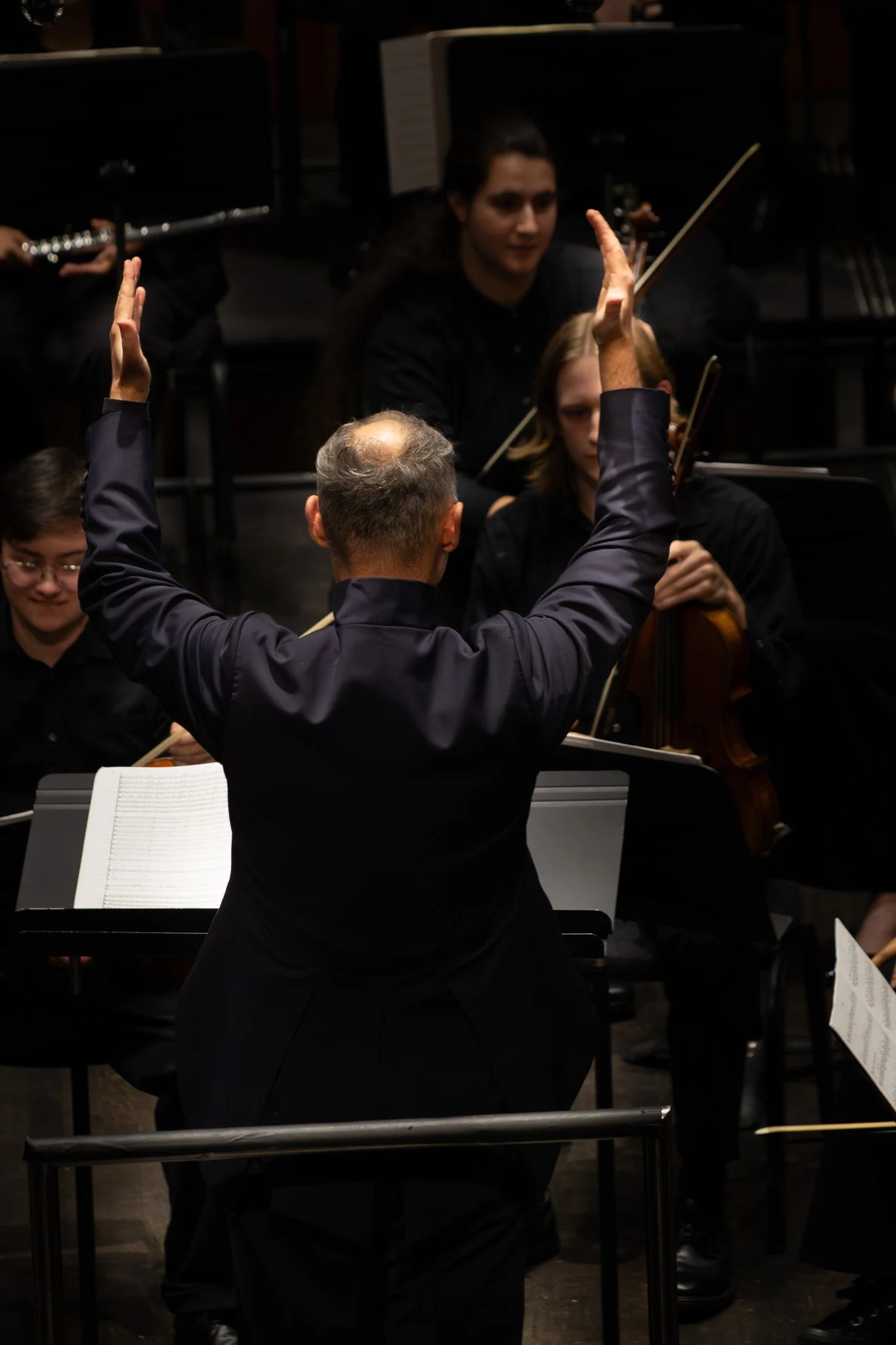 Orchestra conductor leading a group of musicians in a dark concert hall, with some musicians playing string and wind instruments.