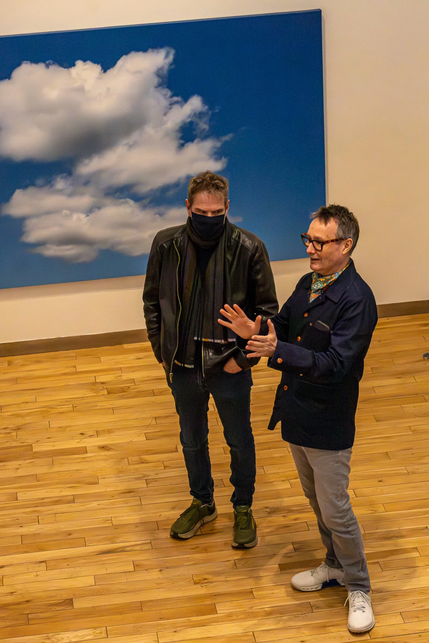 Two men standing and talking inside a room with wooden floors and a large framed photograph of clouds on the wall behind them. One man is wearing a black face mask, black leather jacket, and green shoes. The other man has glasses, a dark jacket, a co